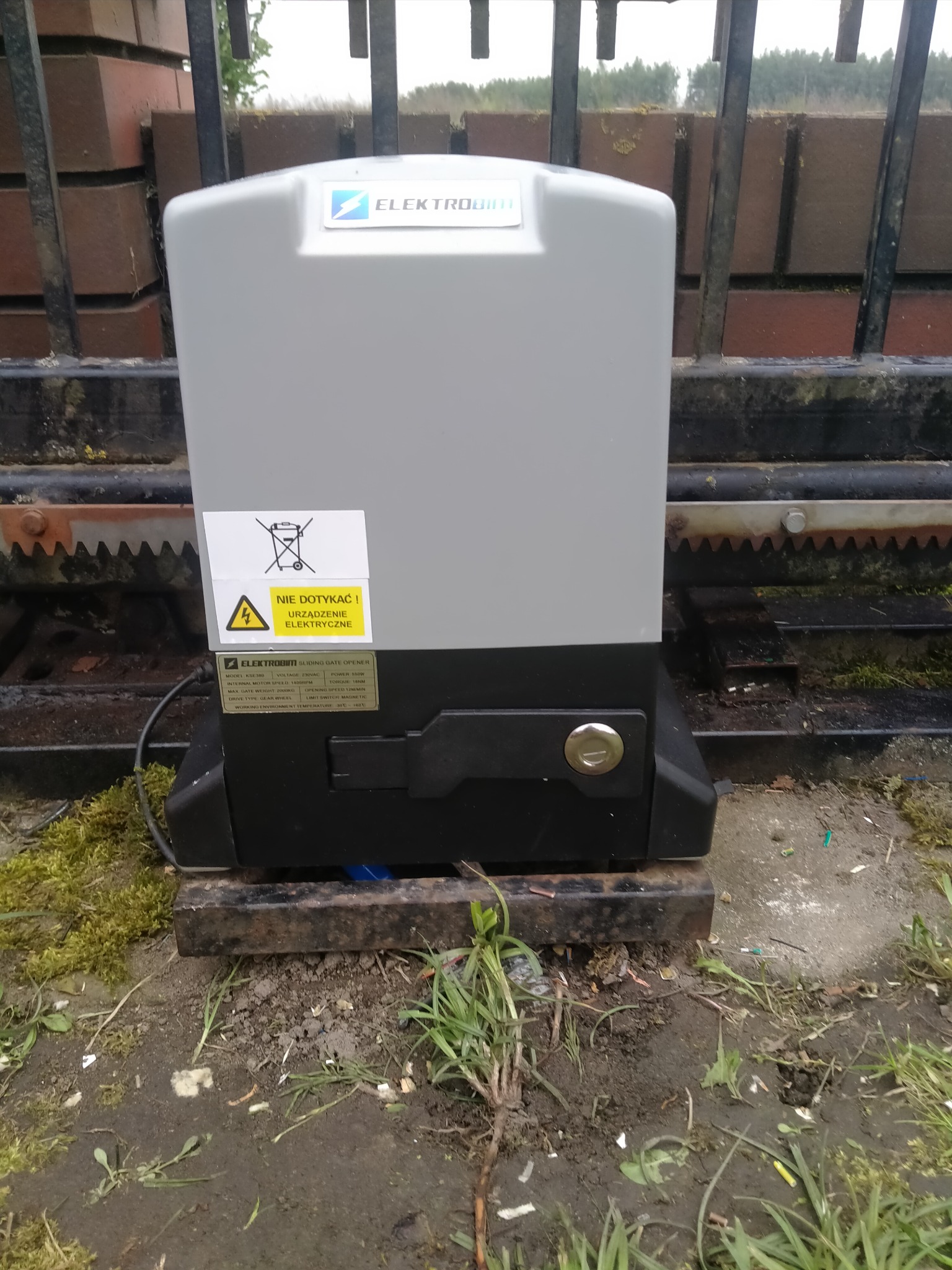 Szary napęd do bramy przesuwnej Elektrobim zamontowany na metalowej podstawie, widoczna zębatka i ostrzeżenie o urządzeniu elektrycznym.