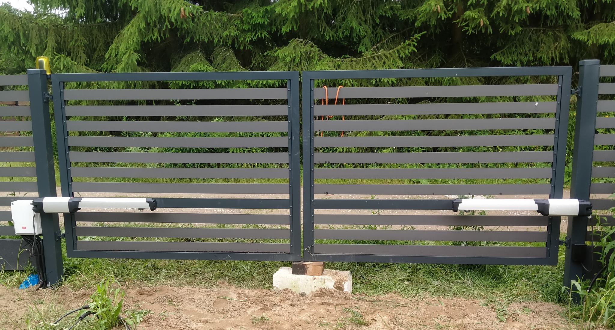 Nowoczesna, pozioma brama wjazdowa z automatyką, widoczna z bliska na tle zieleni. Napędy bramy zamontowane po obu stronach. Brama w kolorze szarym.