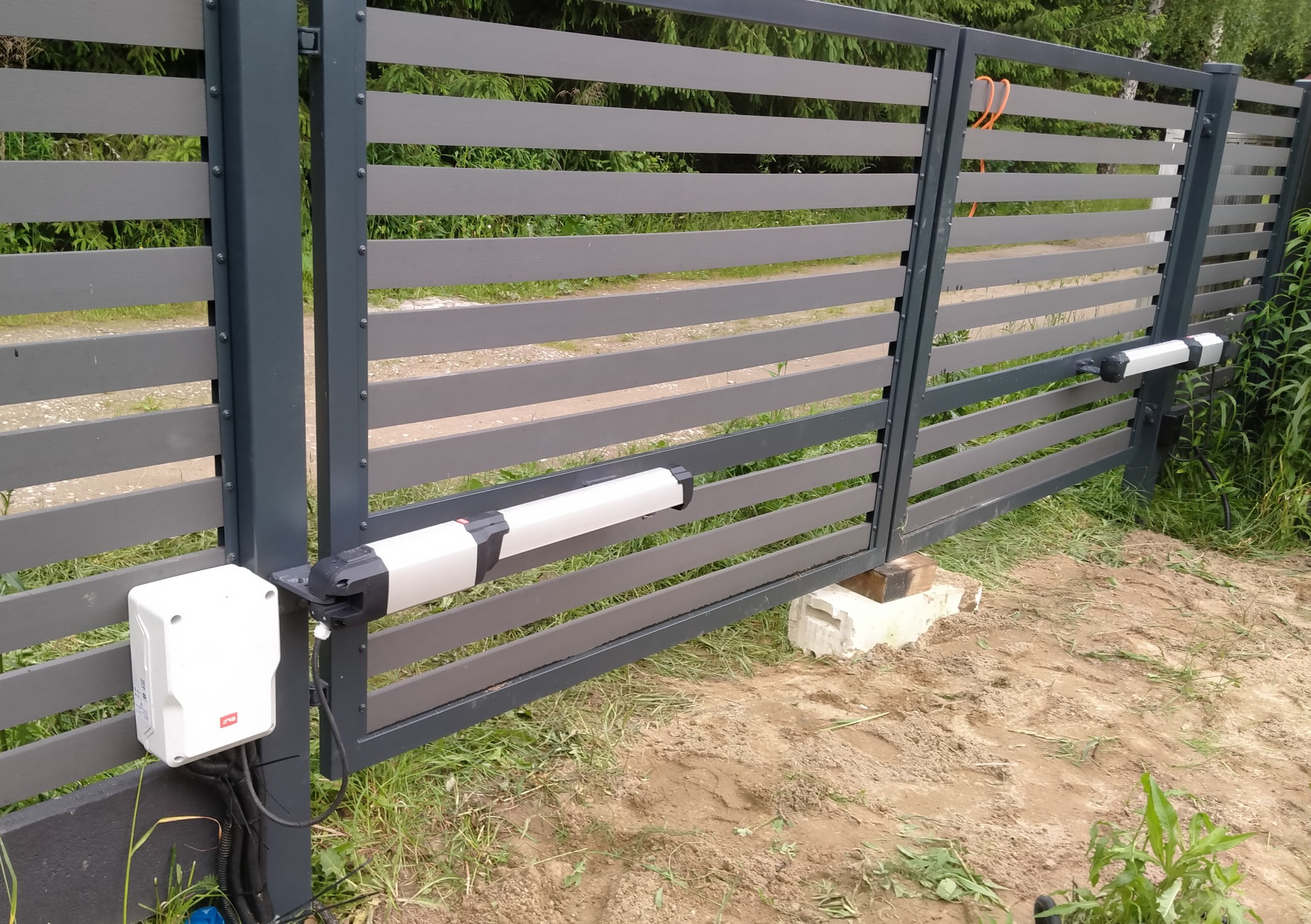 Szara brama przesuwna z automatyką, widoczne siłowniki i centrala sterująca. Brama z poziomych paneli kompozytowych, tło zielone drzewa.