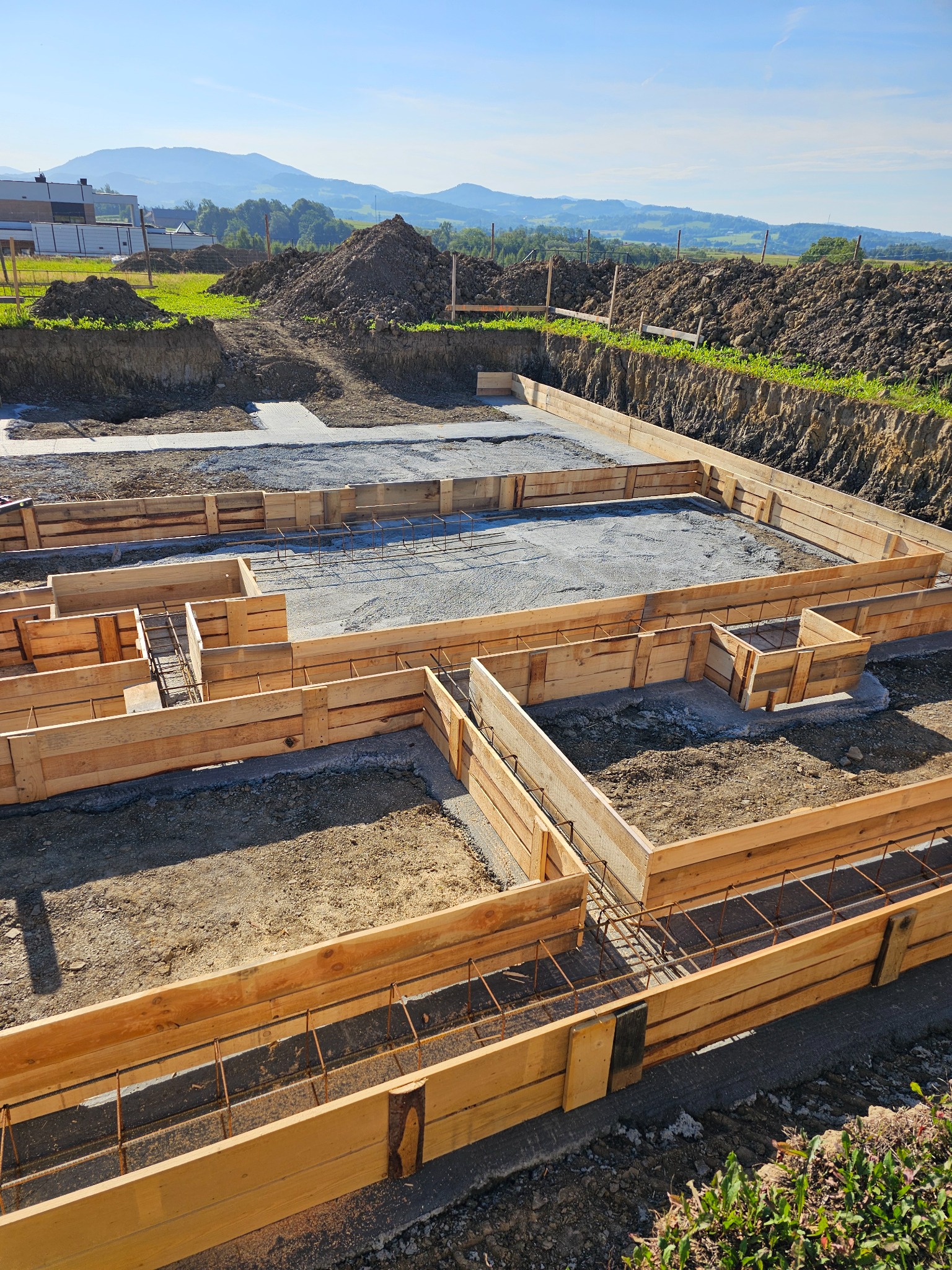 Przygotowanie fundamentów pod budynek: drewniane szalunki zbrojone stalą, częściowo zalane betonem, widoczne usypiska ziemi i krajobraz górski w tle.