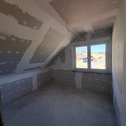 Usługi remontowo wykończeniowe marcin pępek - Poddasze po szpachlowaniu, widoczne łączenia płyt gipsowych. Ściany i sufit przygotowane do malowania, okno z widokiem na zewnątrz. Wnętrze w trakcie wykańczania.