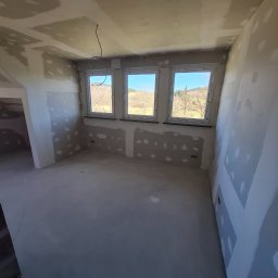 Usługi remontowo wykończeniowe marcin pępek - Pokój po szpachlowaniu, ściany i sufit z widocznymi łatami szpachli, trzy okna z widokiem na zewnątrz, przygotowany do dalszych prac wykończeniowych.