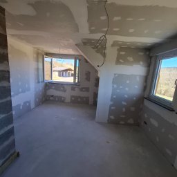 Usługi remontowo wykończeniowe marcin pępek - Wnętrze pokoju w trakcie prac wykończeniowych: ściany z płyt gipsowo-kartonowych, częściowo wyszpachlowane, widoczne okna z widokiem na zewnątrz.