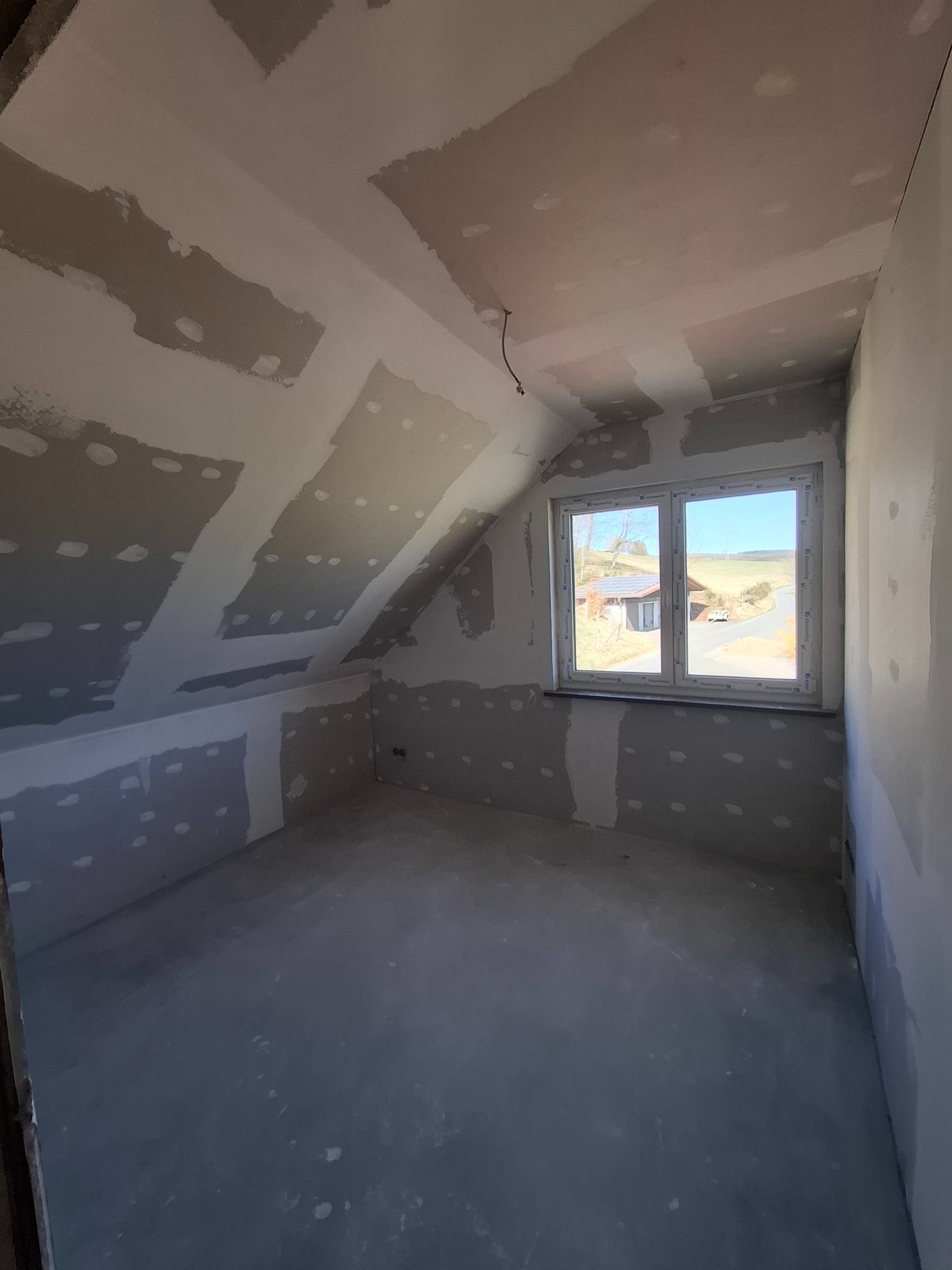 Poddasze po szpachlowaniu, widoczne łączenia płyt gipsowych. Ściany i sufit przygotowane do malowania, okno z widokiem na zewnątrz. Wnętrze w trakcie wykańczania.