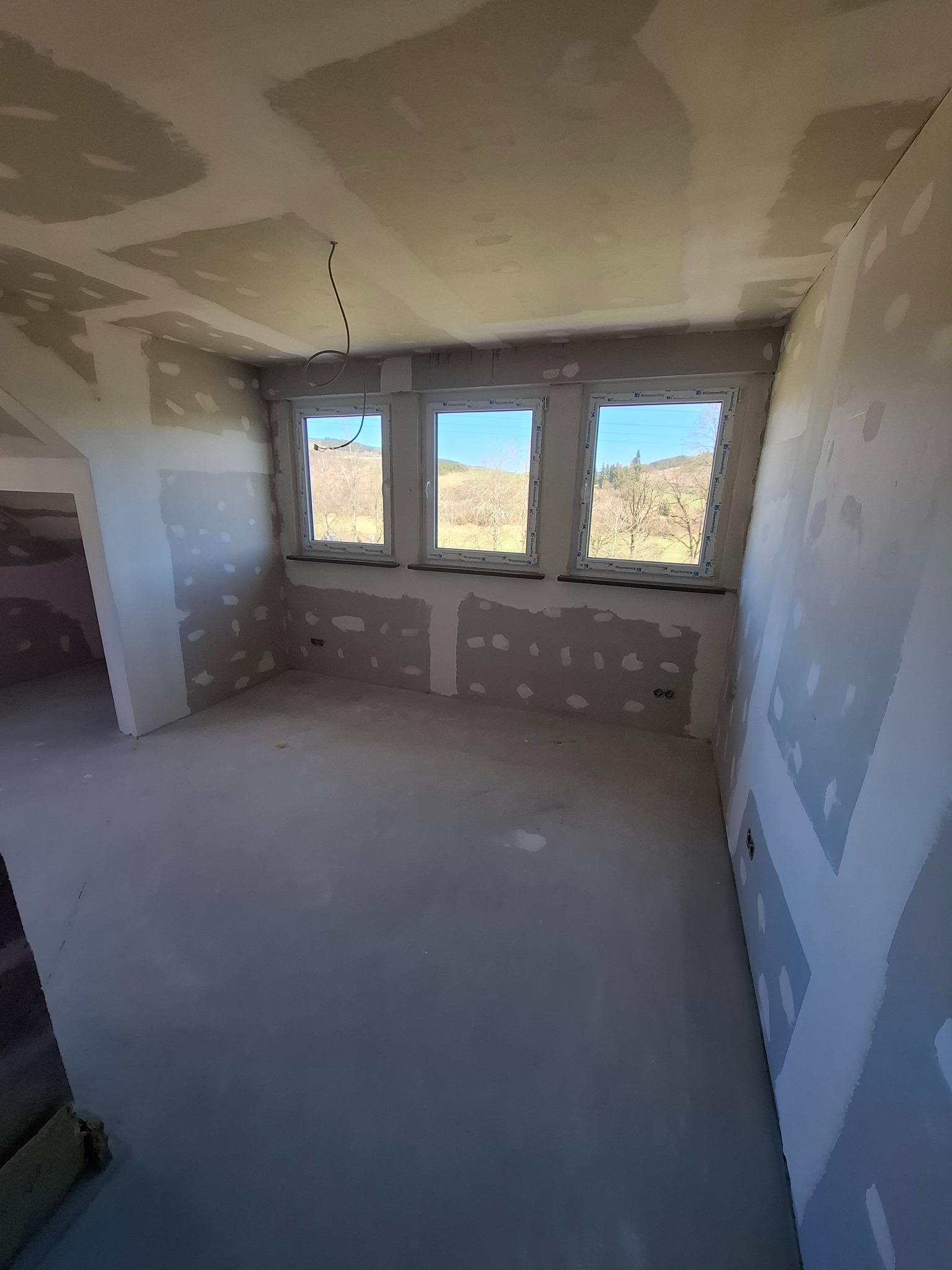 Pokój po szpachlowaniu, ściany i sufit z widocznymi łatami szpachli, trzy okna z widokiem na zewnątrz, przygotowany do dalszych prac wykończeniowych.