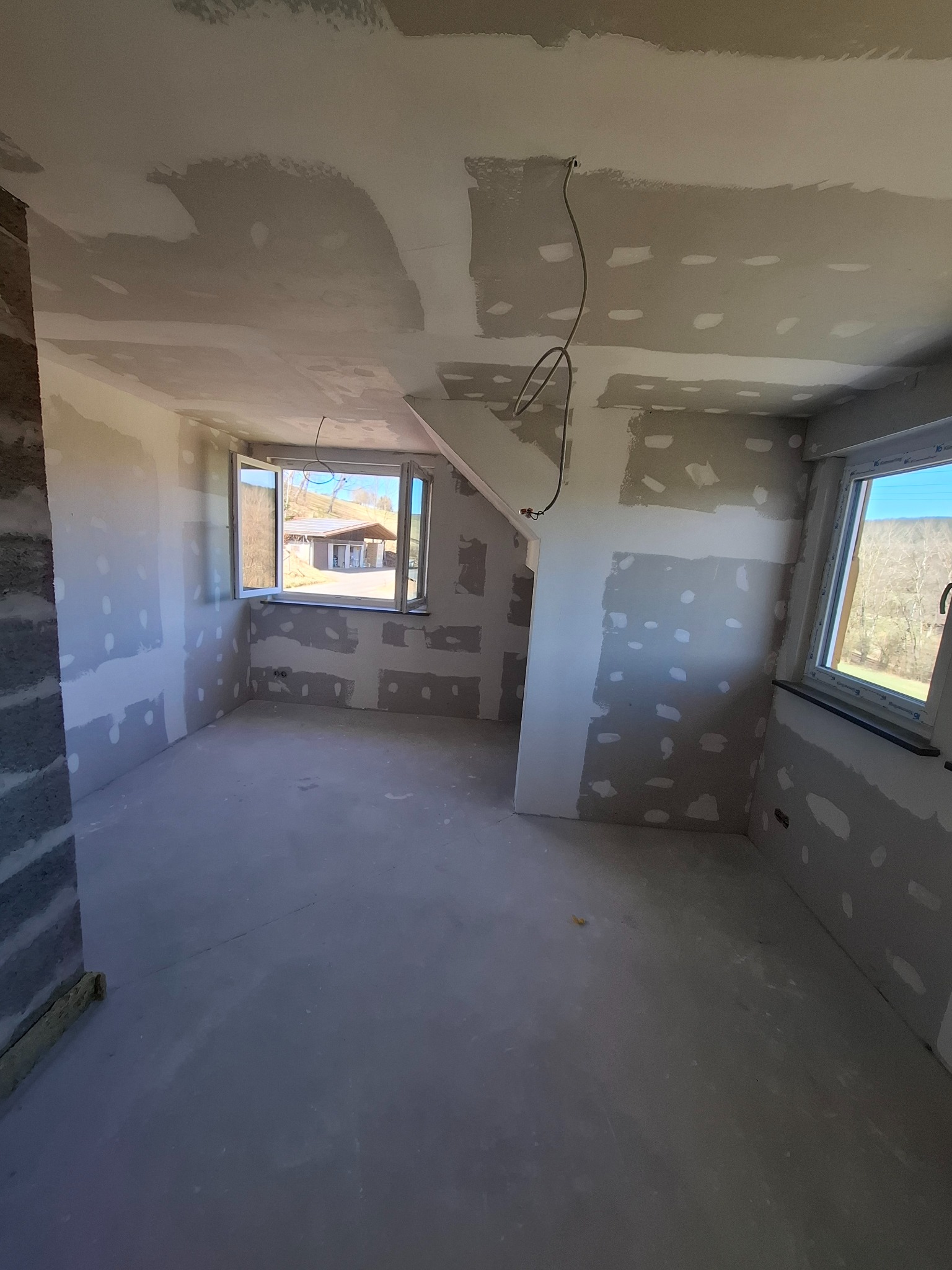 Wnętrze pokoju w trakcie prac wykończeniowych: ściany z płyt gipsowo-kartonowych, częściowo wyszpachlowane, widoczne okna z widokiem na zewnątrz.