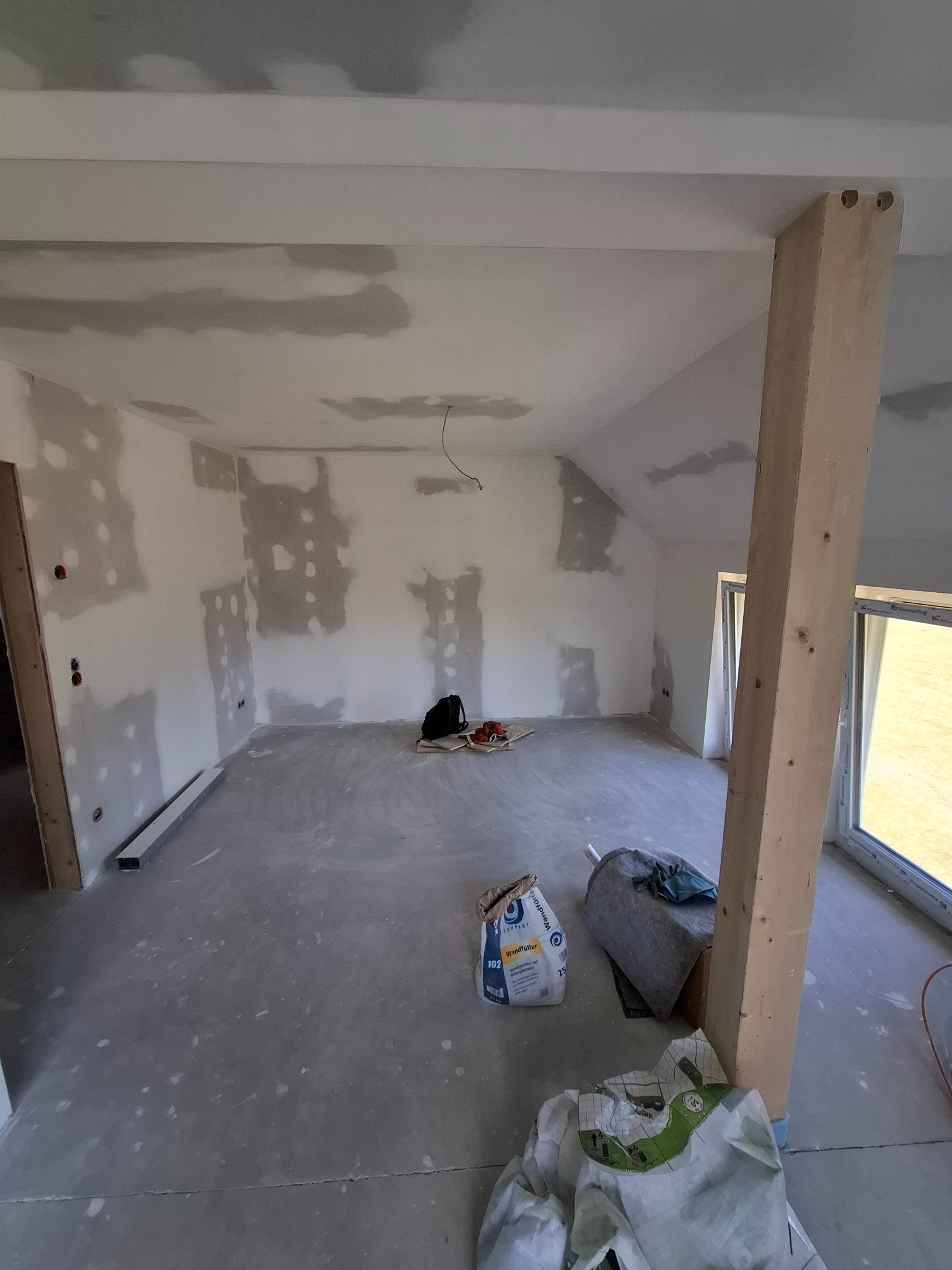 Wnętrze w trakcie prac wykończeniowych: ściany i sufit po szpachlowaniu, widoczne worki z materiałami budowlanymi, drewniany słup konstrukcyjny i okno z widokiem na zewnątrz.