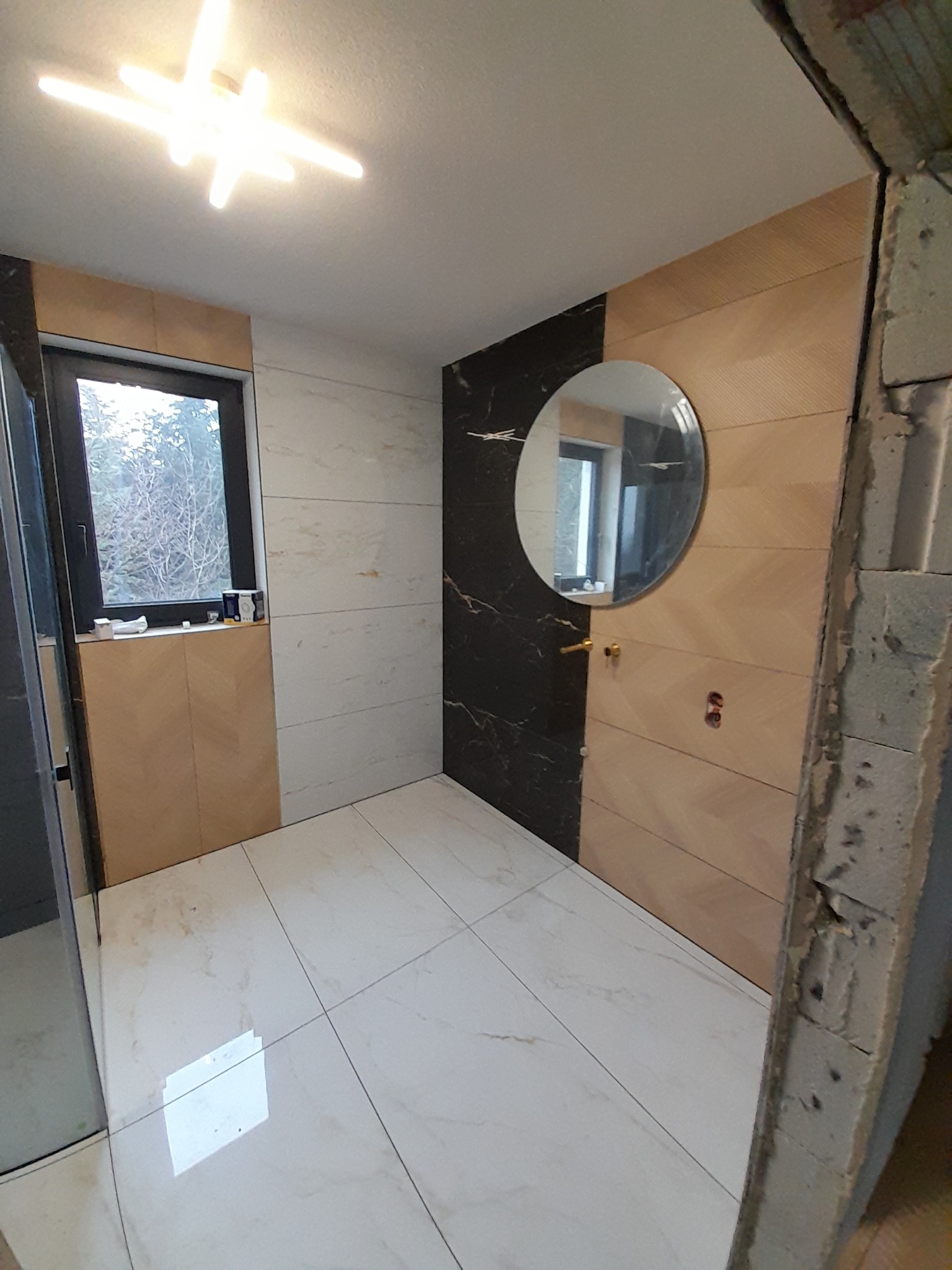 Wykończona łazienka z marmurowymi płytkami, okrągłym lustrem i nowoczesnym oświetleniem. Widoczny fragment surowej ściany, sugerujący trwający remont.