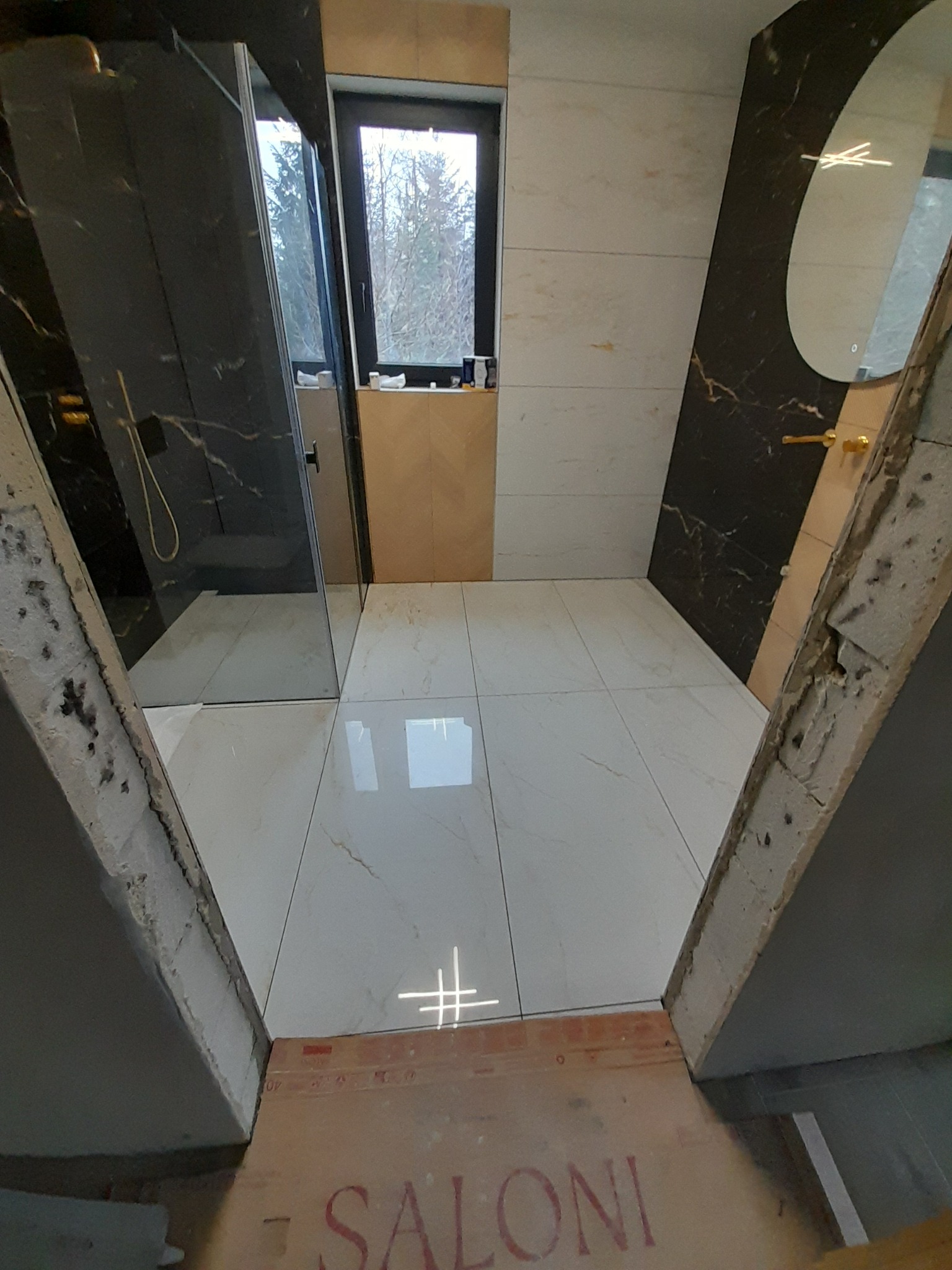 Nowoczesna łazienka w trakcie wykańczania: białe płytki podłogowe, kabina prysznicowa z czarnymi elementami, owalne lustro z podświetleniem. Widoczne surowe wykończenia.