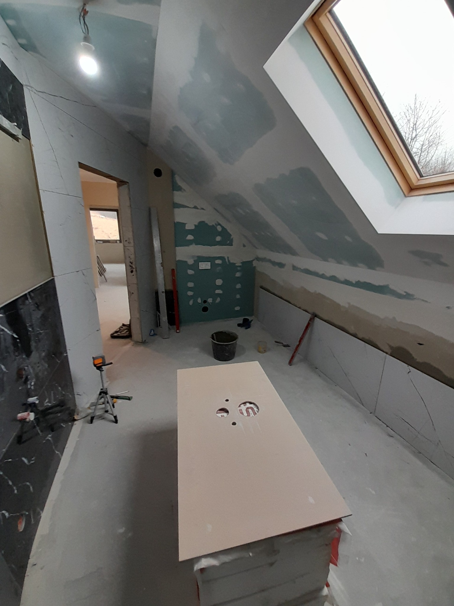 Poddasze w trakcie wykańczania: płyty gipsowe na skosach, okno dachowe, przygotowana płyta pod umywalkę. Widoczne narzędzia i materiały budowlane.
