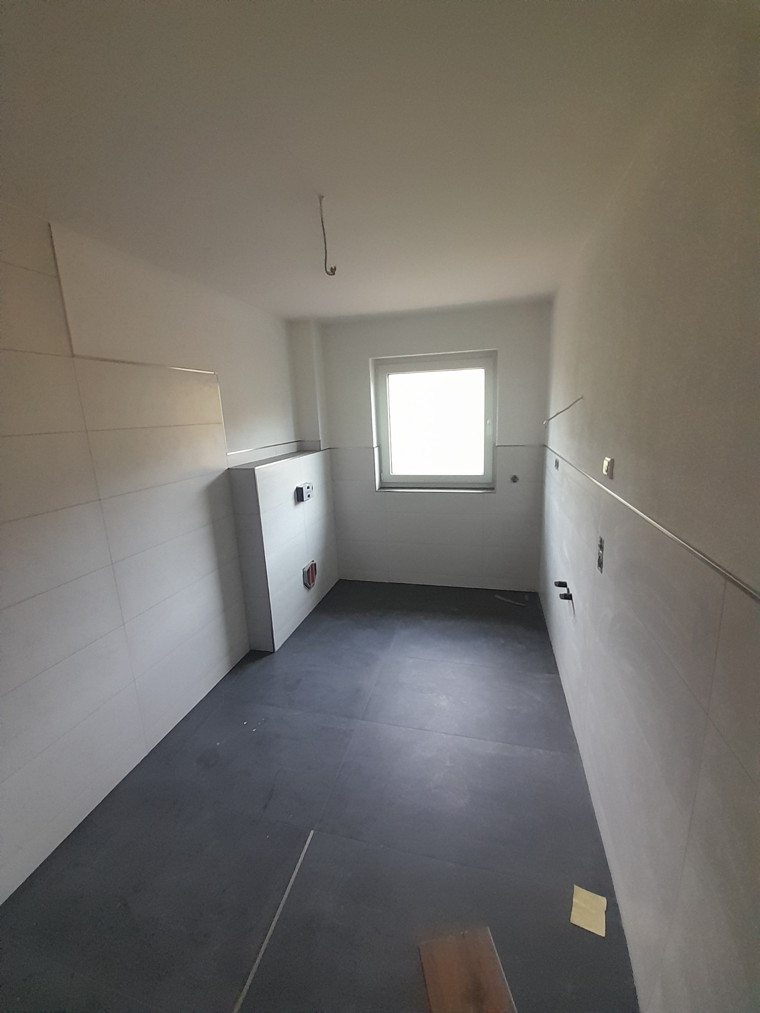 Wnętrze łazienki po remoncie: szare płytki podłogowe, białe płytki ścienne z listwą, okno z białym parapetem, zabudowa geberitu. Widok z góry, neutralne oświetlenie.