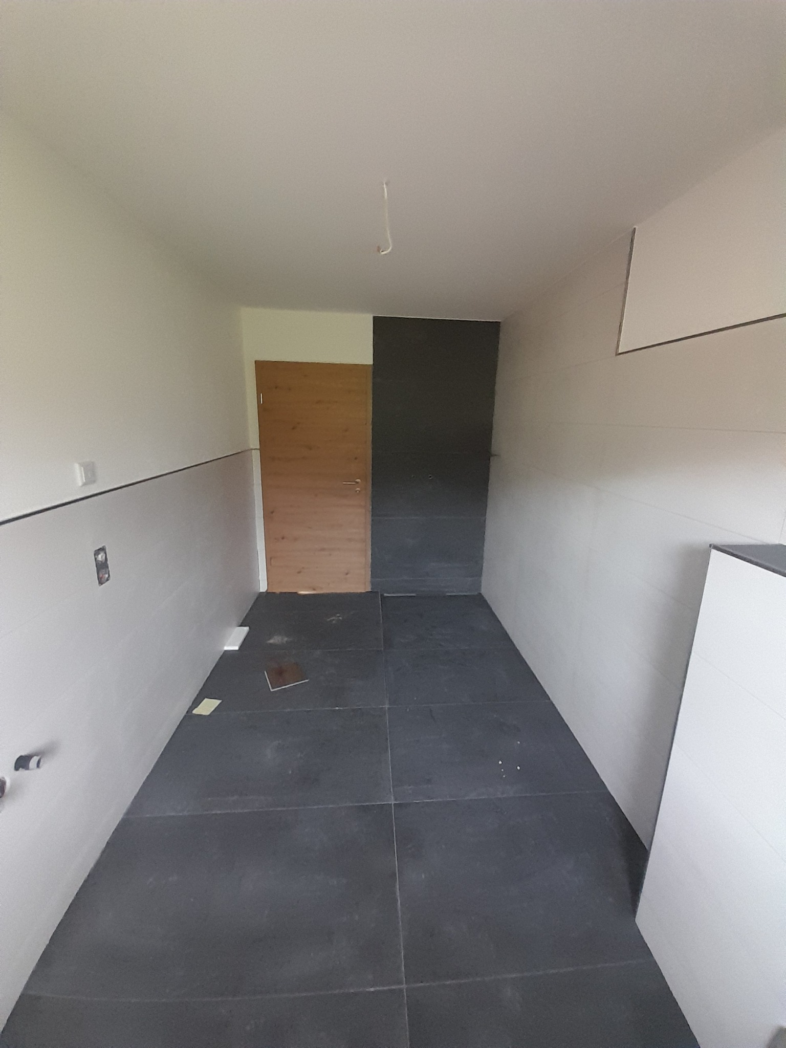 Wykończona łazienka z ciemną podłogą i jasnymi ścianami. Widoczna drewniana ościeżnica drzwiowa i fragment zabudowy. Minimalistyczny styl, surowe wykończenie.