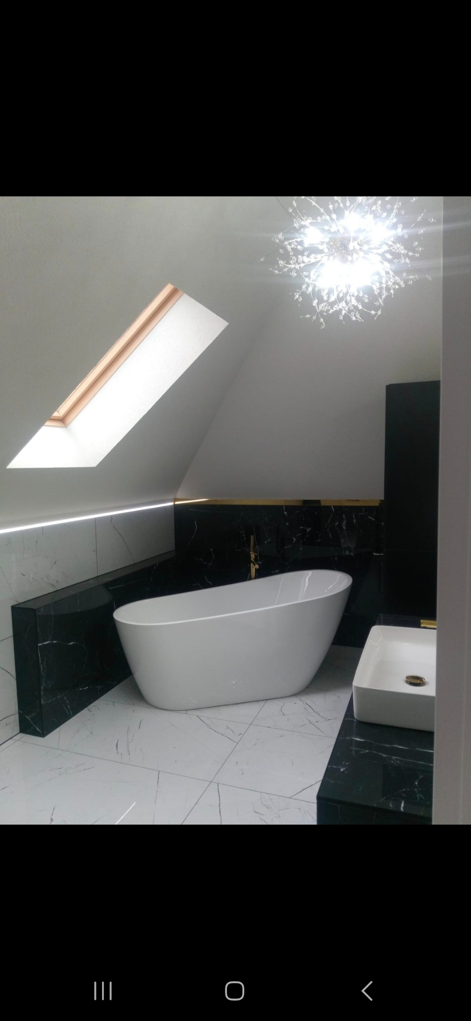 Nowoczesna łazienka na poddaszu z wolnostojącą wanną, białą armaturą i czarnymi elementami z marmuru. Okno dachowe doświetla wnętrze.