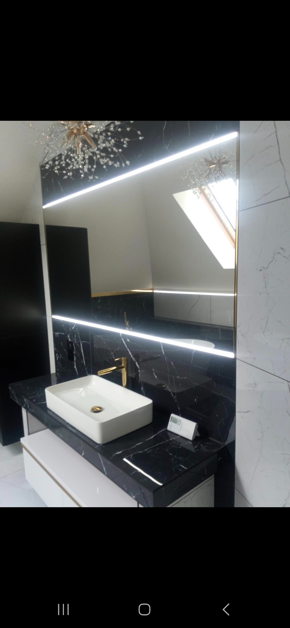 Nowoczesna łazienka z czarnym marmurem, białym umywalką i złotą armaturą. Lustro z podświetleniem LED i dekoracyjnym żyrandolem. Elegancki design.