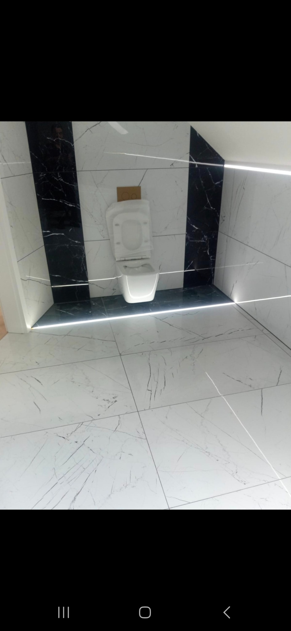 Nowoczesna łazienka po remoncie: białe płytki z marmurowym wzorem, czarne akcenty, podświetlenie LED i podwieszana toaleta z deską w folii ochronnej.