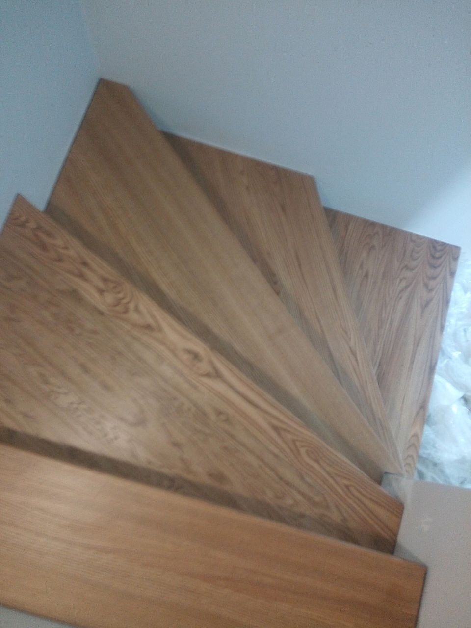 Fragment drewnianych schodów z widocznym usłojeniem drewna, zabezpieczonych folią ochronną na dole, ujęcie z góry.