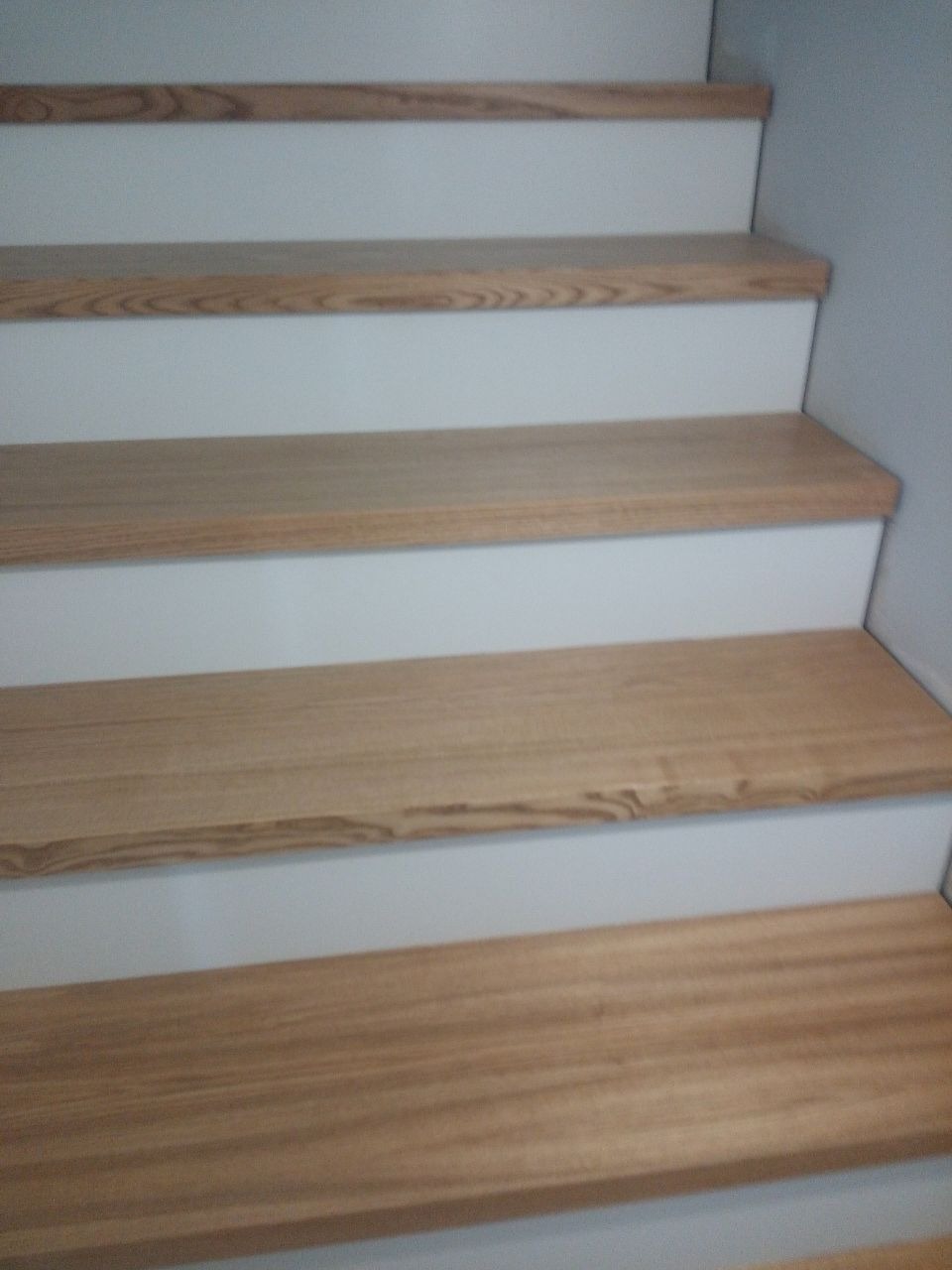 Stopnie schodów z jasnego drewna dębowego, wykończone białymi podstopnicami i boczną ścianą, widoczne z dołu pod kątem.