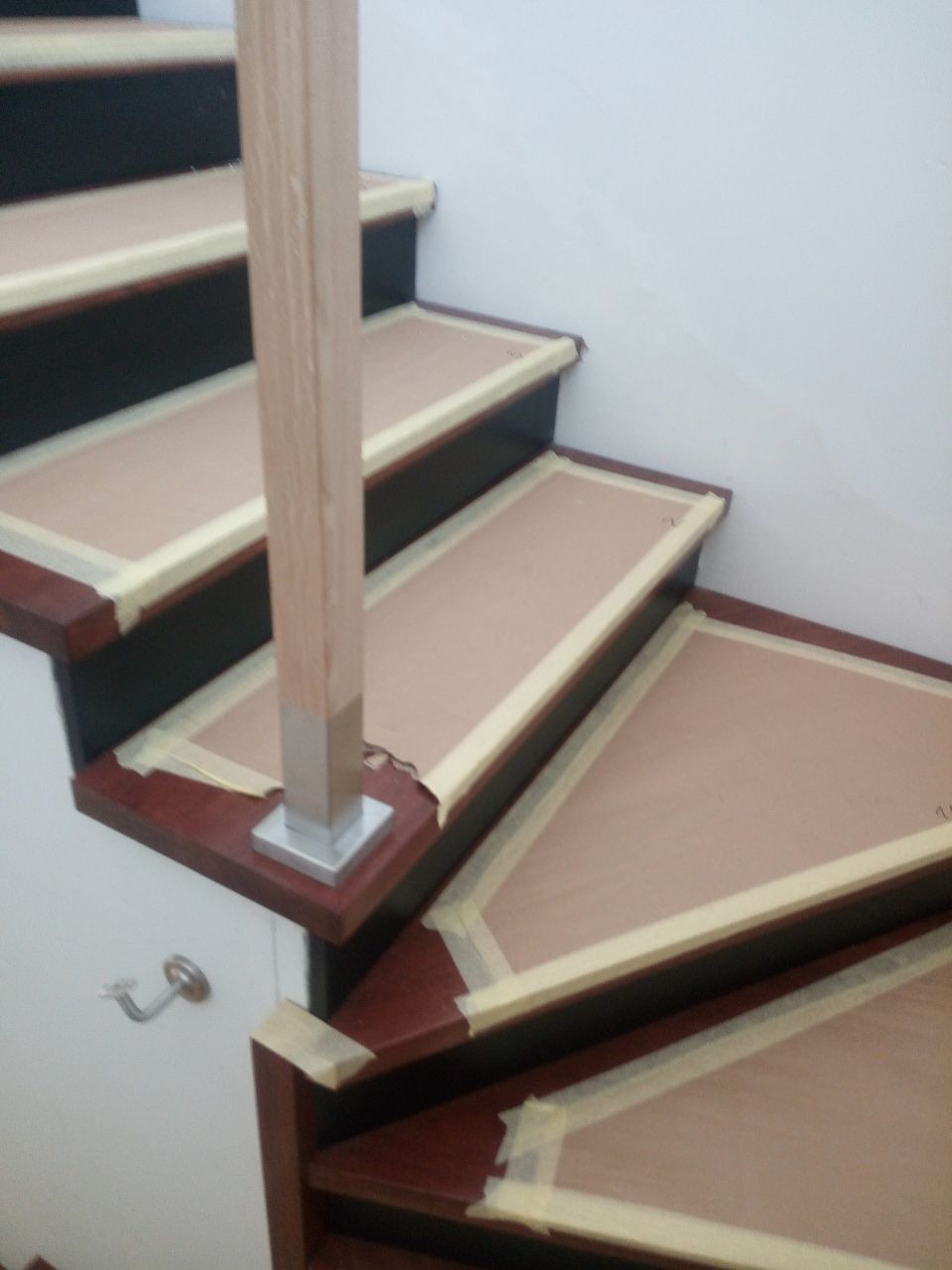 Fragment drewnianych schodów z czarnymi podstopnicami, zabezpieczonych taśmą malarską przed malowaniem stopni, widoczny fragment poręczy z metalową podstawą