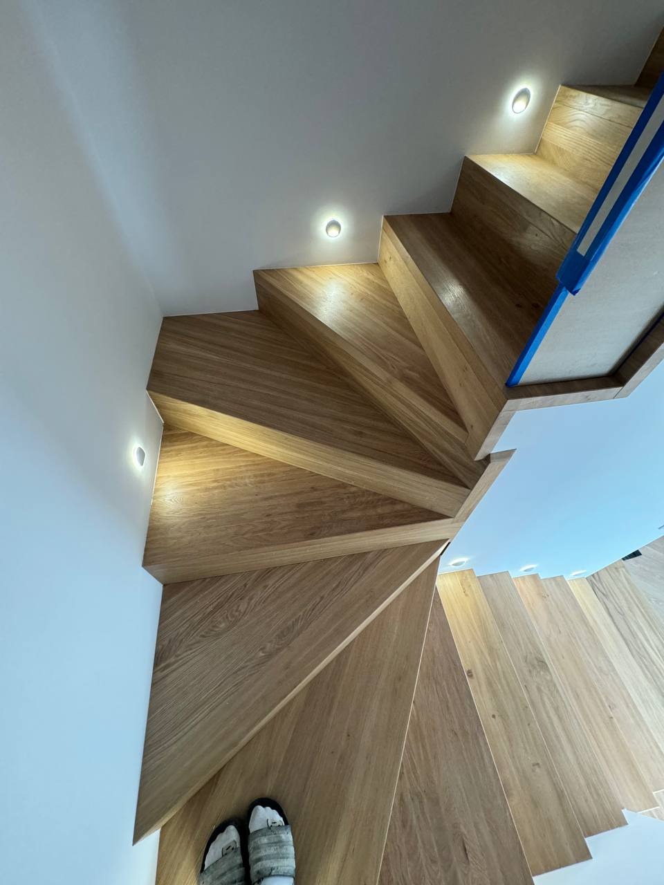 Drewniane schody kręcone z oświetleniem punktowym w ścianie, widok z góry, na dole schodów leżą kapcie.