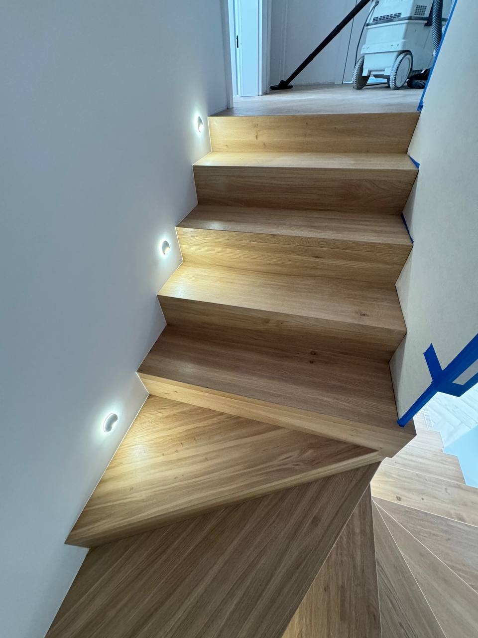 Nowoczesne drewniane schody z oświetleniem LED w ścianie, widoczny odkurzacz przemysłowy w tle, zabezpieczone taśmą malarską.