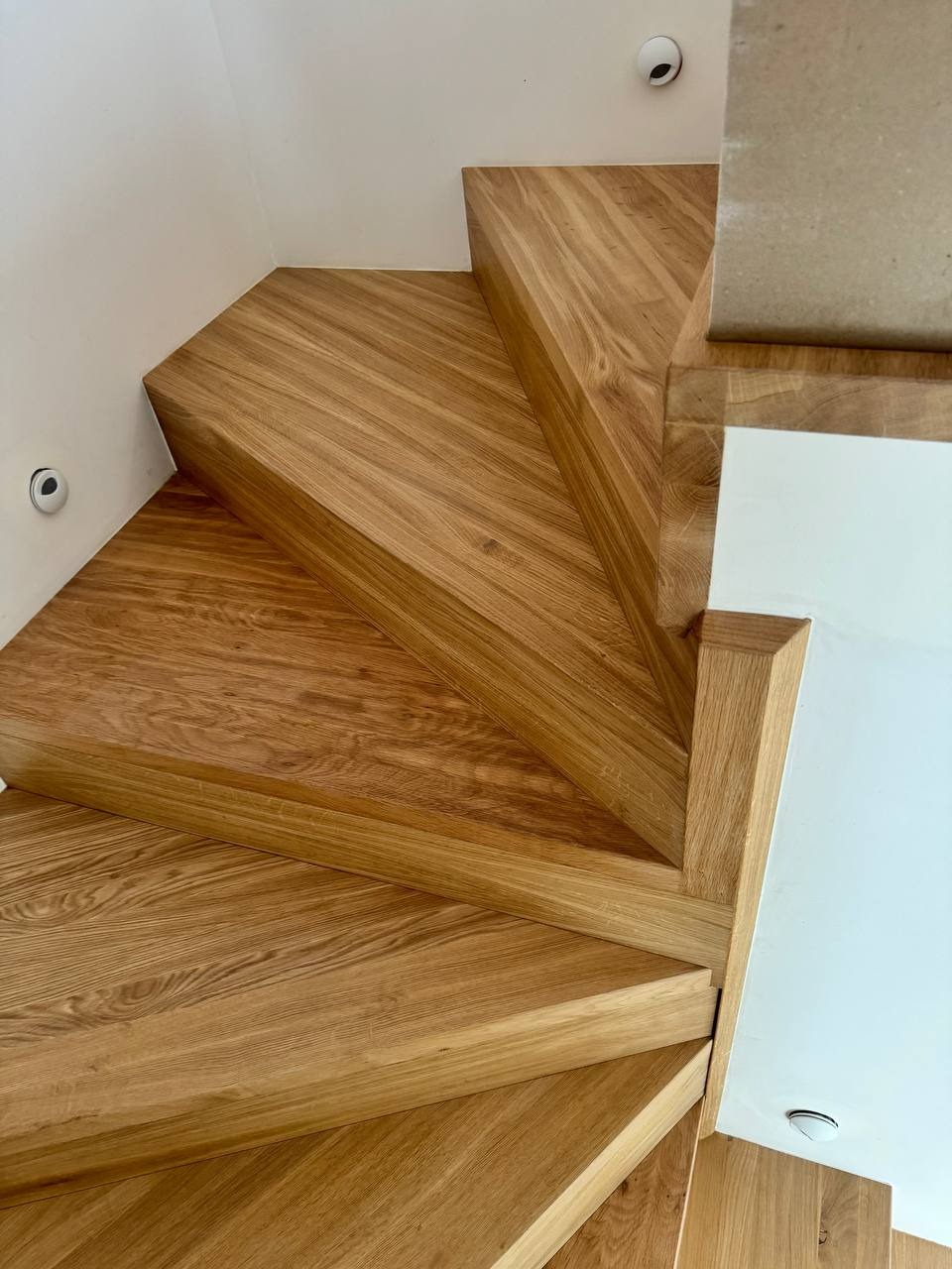 Jasne, dębowe stopnie schodów z wyraźnym usłojeniem, widok z góry, minimalistyczne oświetlenie punktowe w białym suficie.