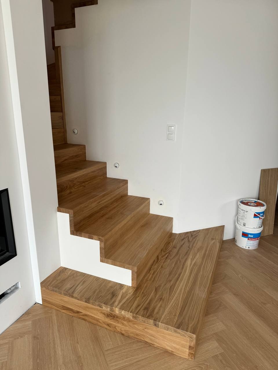 Stopnie z jasnego drewna, z białymi podstopnicami, oświetlenie punktowe w ścianie, parkiet ułożony w jodełkę, puszki z farbą w tle.