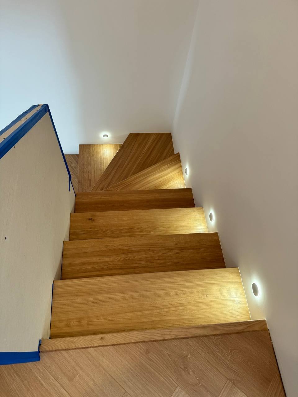 Nowoczesne, drewniane schody z podświetleniem LED w ścianie, wykończone parkietem w jodełkę, zabezpieczone taśmą malarską przed zabrudzeniem.