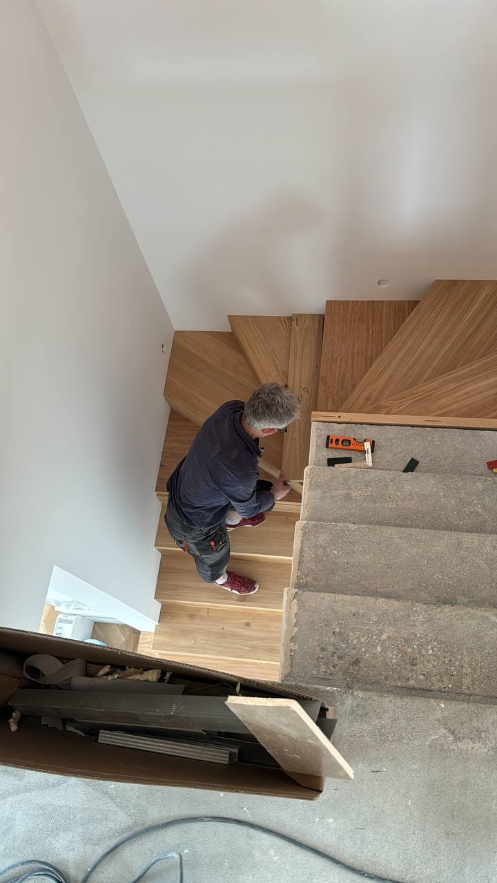Montaż drewnianych schodów z widocznym stolarzem docinającym element wykończeniowy, surowe betonowe stopnie obok.