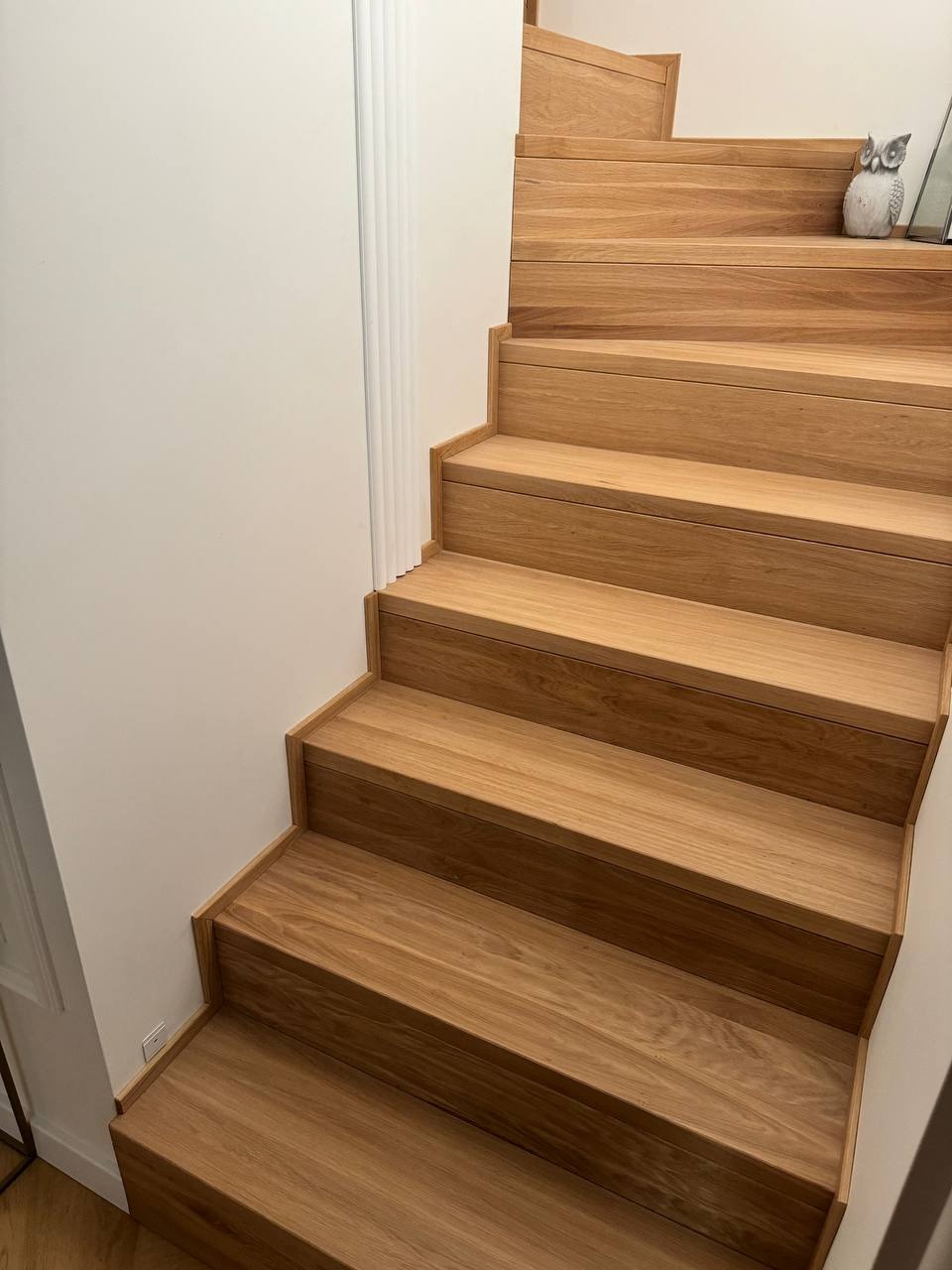 Jasne, drewniane stopnie schodów z widocznym usłojeniem, przylegające do białej ściany z dekoracyjną listwą. Na górnym stopniu dekoracyjna figurka sowy.