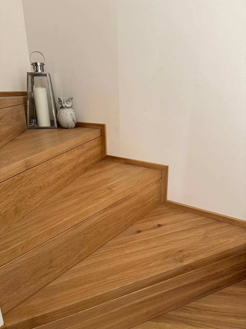 Jasne, drewniane stopnie schodów z dekoracyjną latarenką i figurką sowy na górnym stopniu, przy białej ścianie.