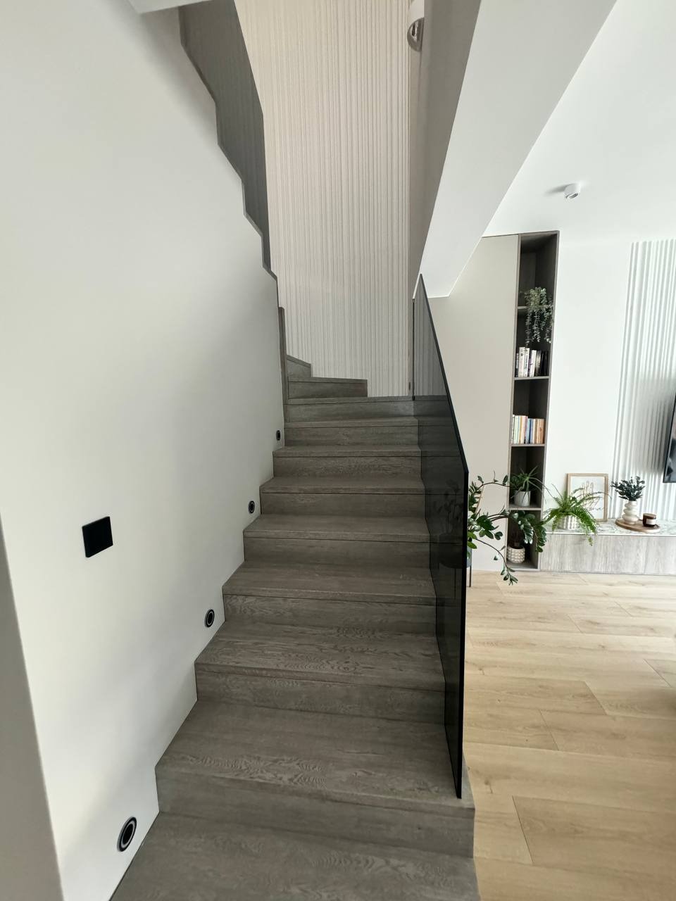 Nowoczesne schody z szarego drewna z czarną szklaną balustradą i punktowym oświetleniem ściennym, prowadzące do jasnego salonu z drewnianą podłogą i roślinami.