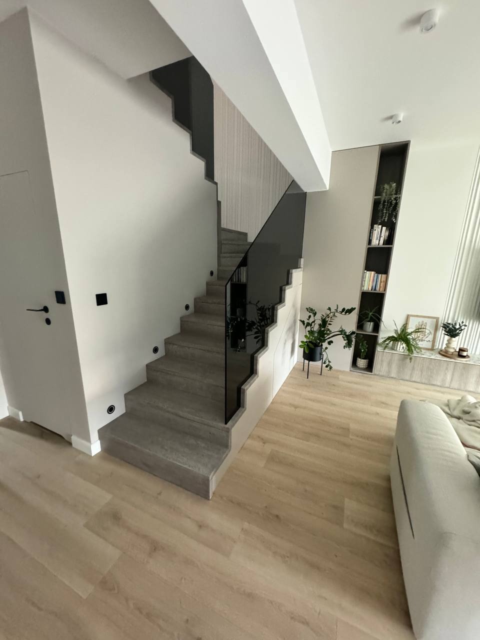 Nowoczesne schody z szarymi stopniami i czarną szklaną balustradą w jasnym wnętrzu z drewnianą podłogą i minimalistycznymi dekoracjami.