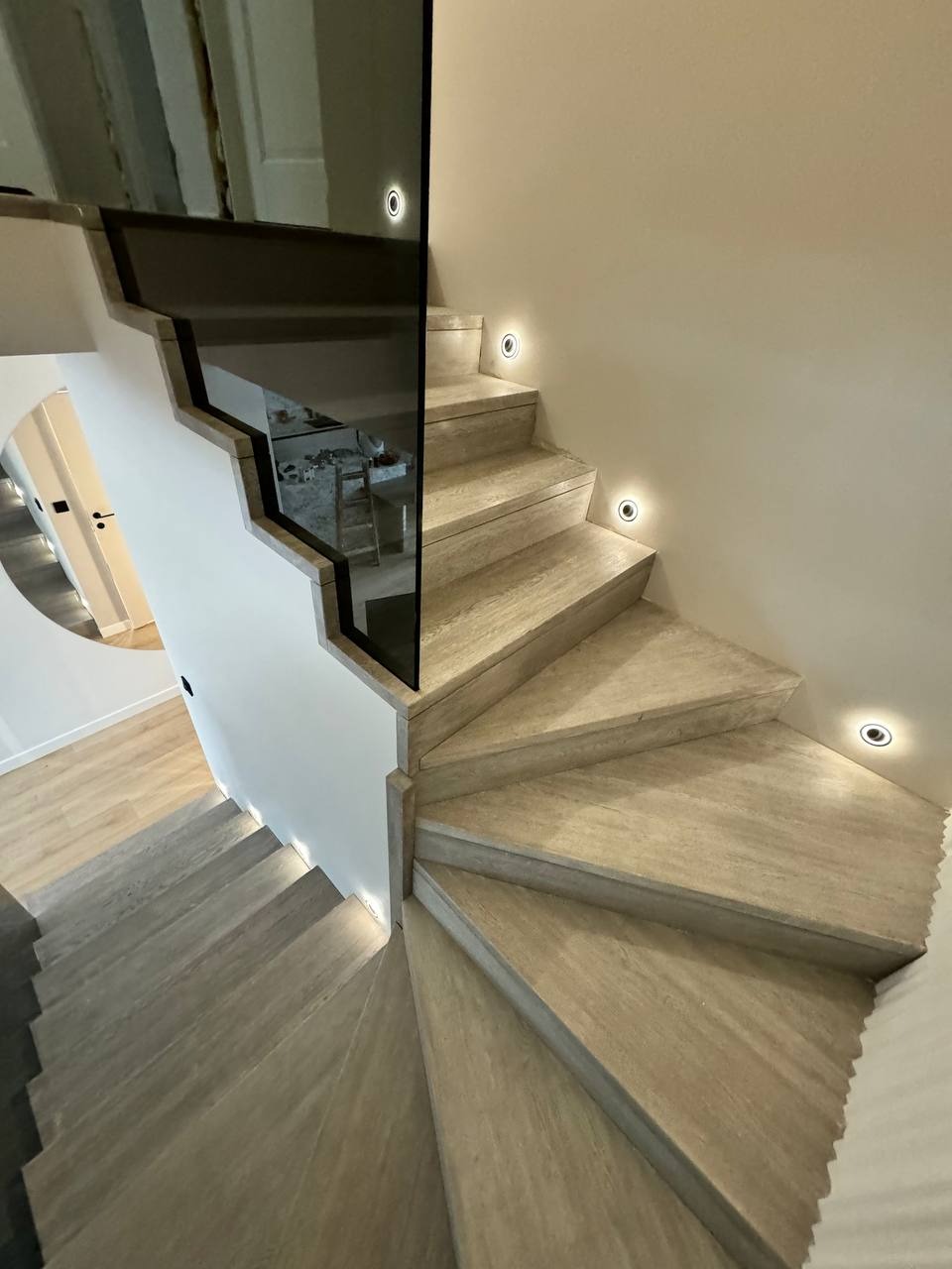 Spiralne schody z jasnego drewna z podświetleniem LED w ścianie i szklaną balustradą odbijającą wnętrze pomieszczenia.