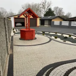 Ułożona mozaika z kostki brukowej w odcieniach szarości i czerni tworząca wzór na podjeździe, z czerwoną studnią w centrum i elementami małej architektury krajobrazu.