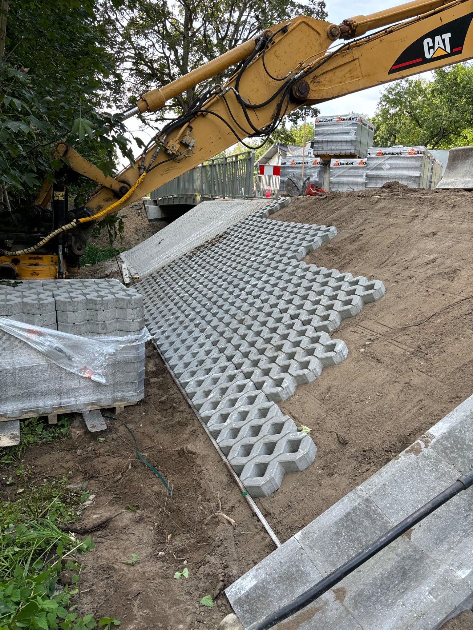 Żółta koparka CAT układa ażurowe płyty betonowe na zboczu, paleta z zapakowanymi płytami obok, widoczne ślady pracy ziemnych.