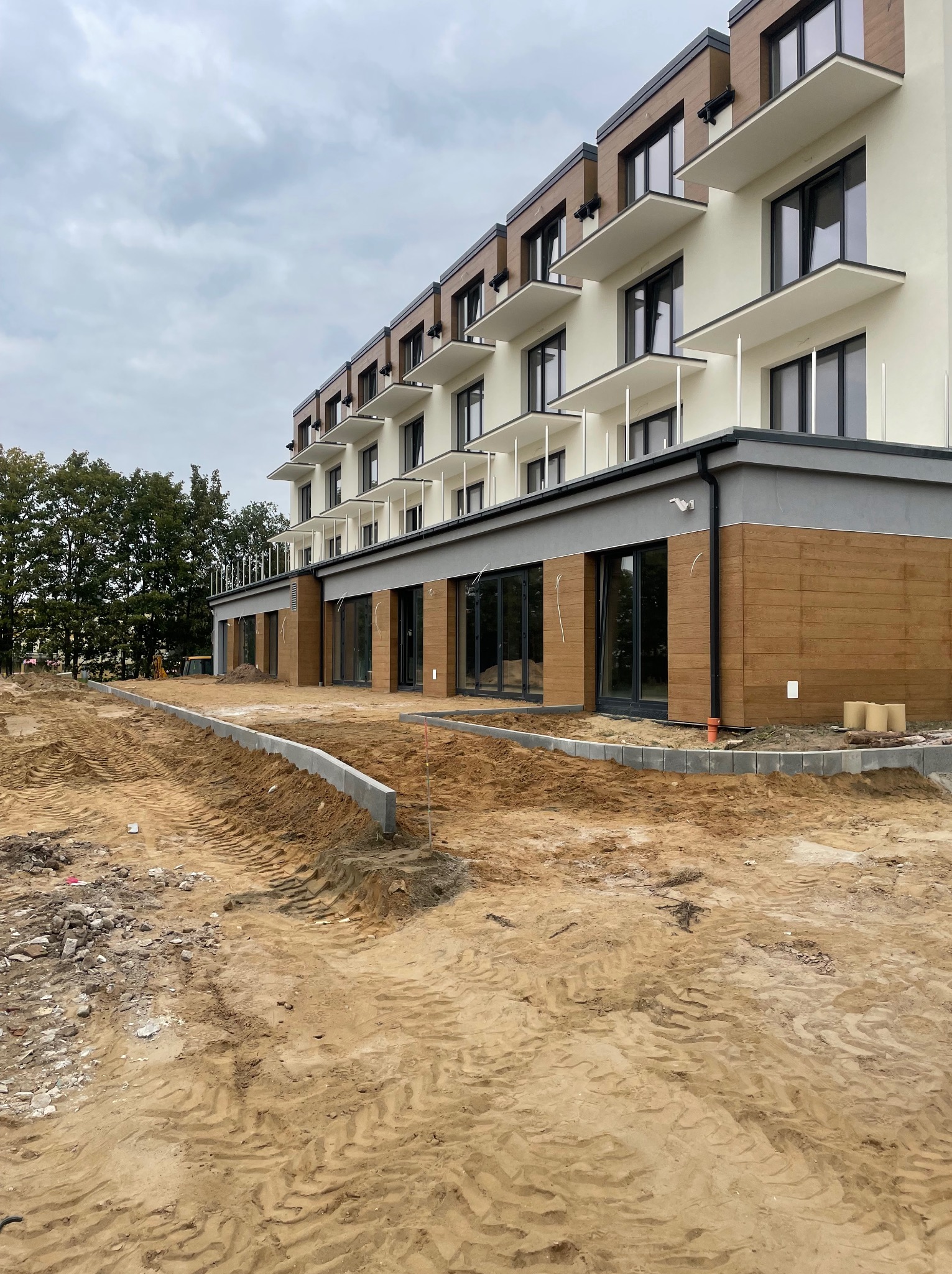 Budynek wielorodzinny w trakcie wykończenia terenu zewnętrznego, widoczny obrys chodnika z kostki, przygotowany teren pod przyszły trawnik, w tle drzewa i pochmurne niebo.