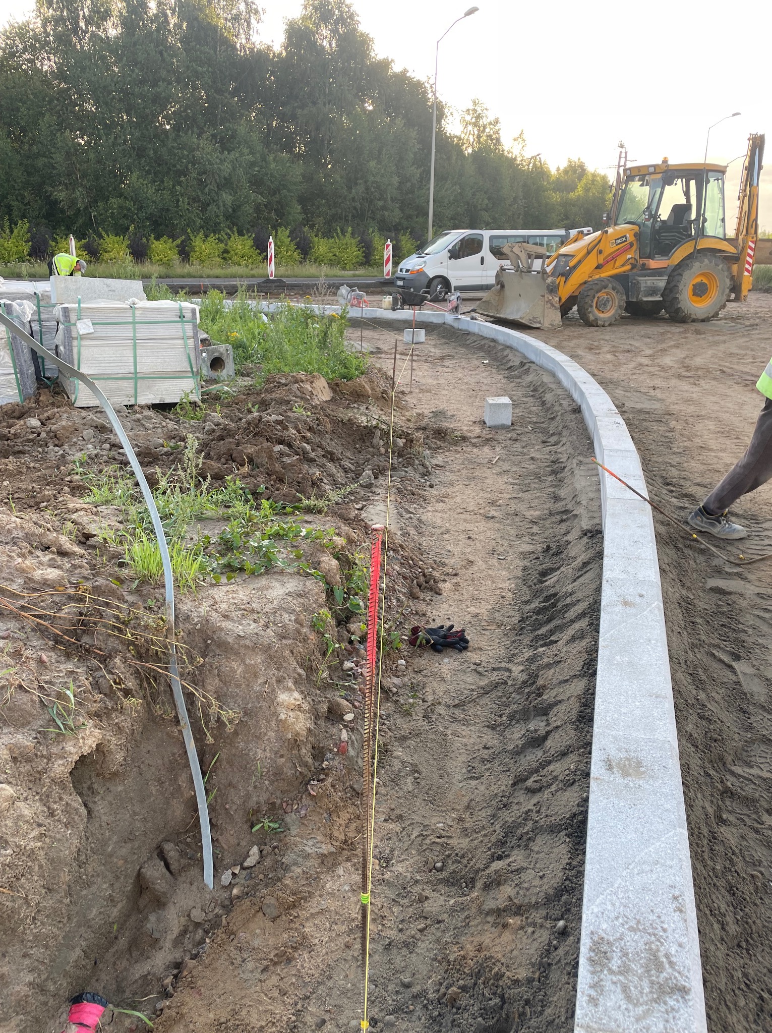 Montaż obrzeży chodnikowych z kostki granitowej na przygotowanym podłożu, widoczny żółty traktor i palety z kostką w tle.