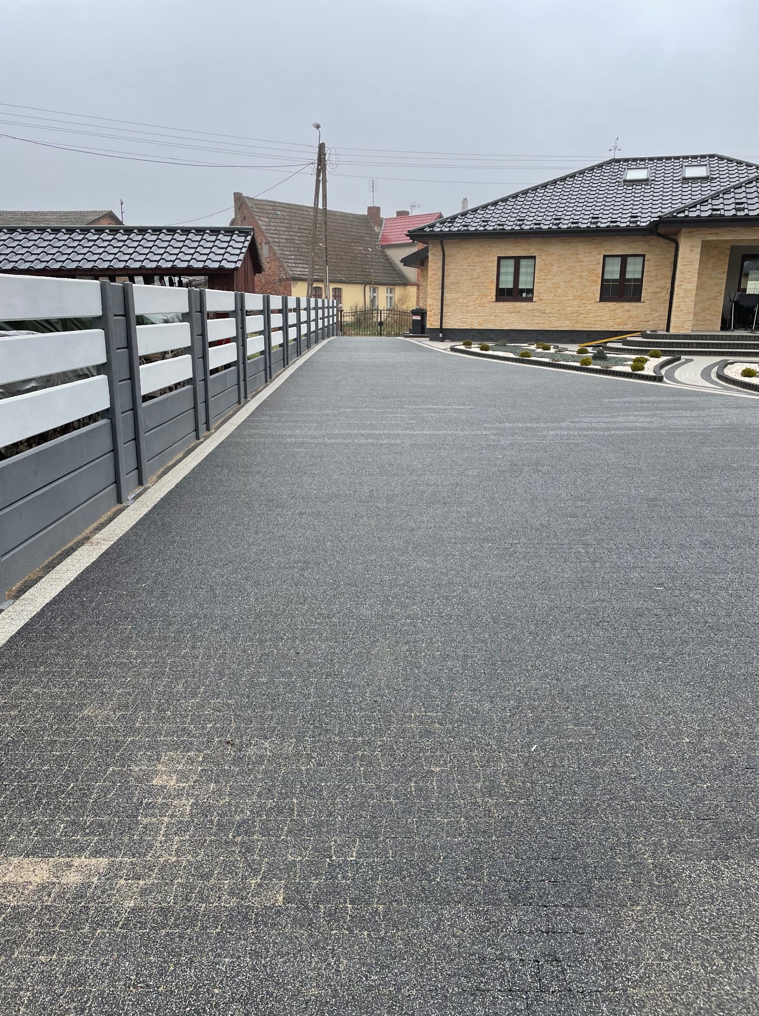 Szeroki, szary podjazd z kostki brukowej prowadzący do domu z beżowej cegły, obok nowoczesne ogrodzenie panelowe w kolorach szarości i bieli, widoczne ślady piasku na powierzchni kostki.