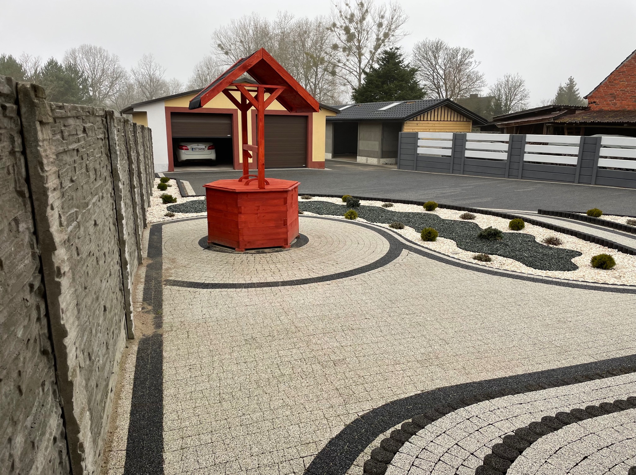 Ułożona mozaika z kostki brukowej w odcieniach szarości i czerni tworząca wzór na podjeździe, z czerwoną studnią w centrum i elementami małej architektury krajobrazu.