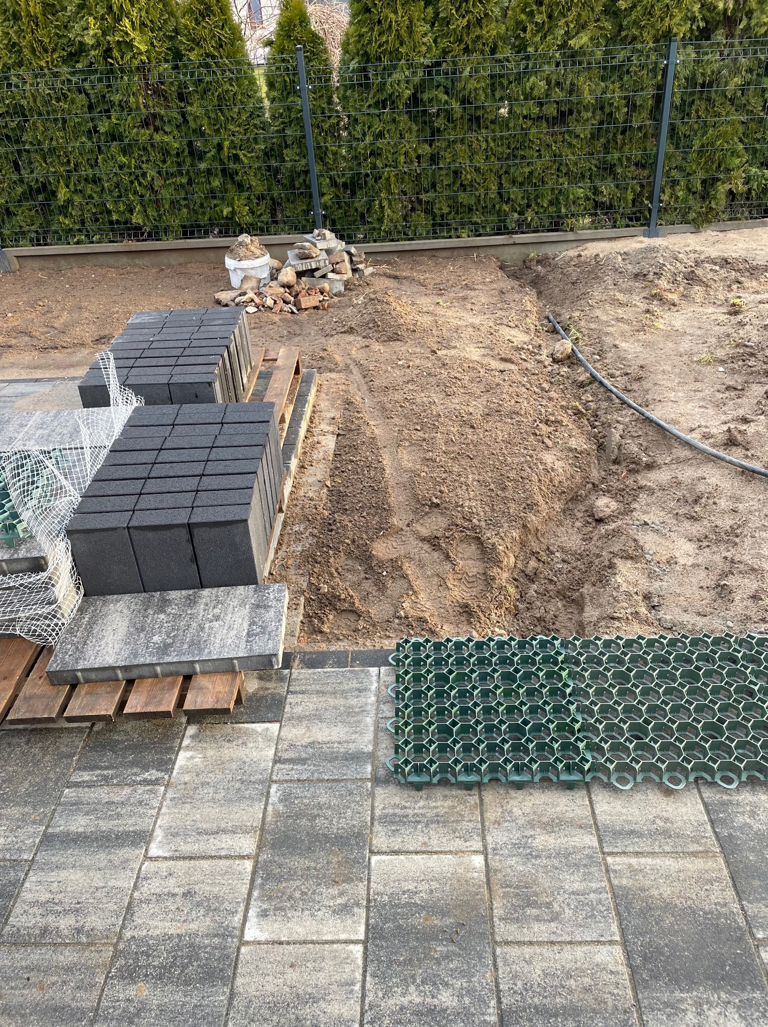 Układanie kostki brukowej: widok na częściowo ułożoną szarą kostkę brukową, paletę z czarną kostką oraz przygotowany teren pod dalszą pracę, z widoczną siatką wzmacniającą podłoże.