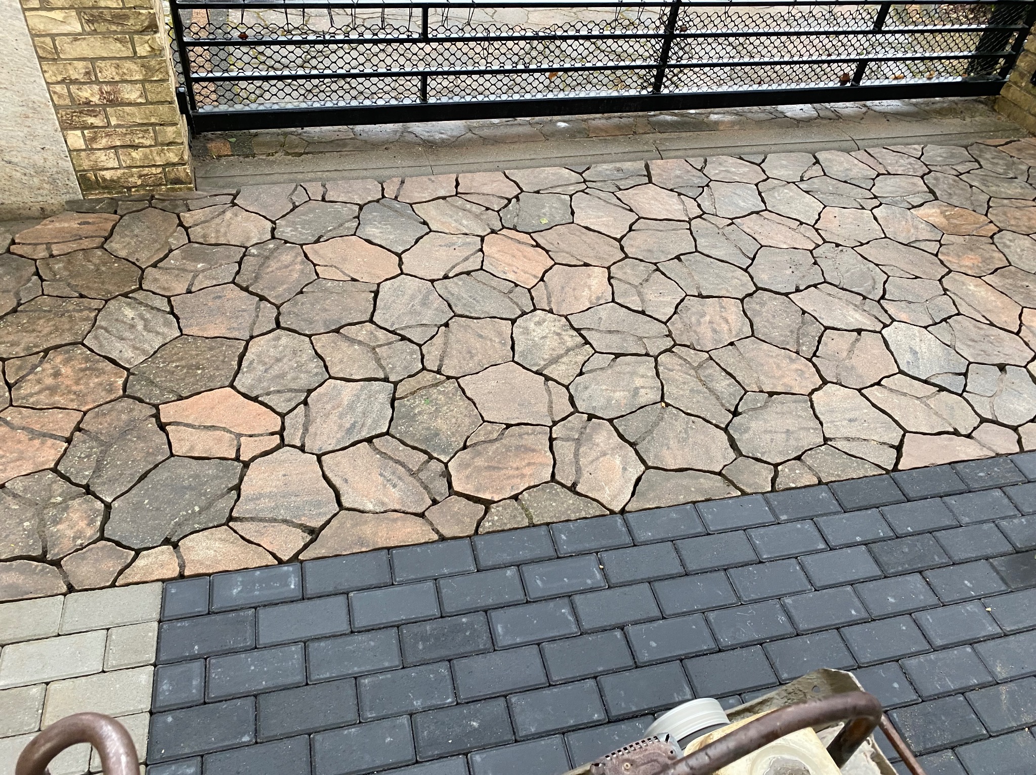 Ułożona nawierzchnia z nieregularnych, brązowo-szarych kamieni obok fragmentu nowej, ciemnoszarej kostki brukowej i bramy wjazdowej.