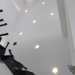 Wysoka ściana z zamontowanymi okrągłymi lampami wpuszczanymi, widok z dołu, obok czarne schody i wisząca lampa z białych kostek.