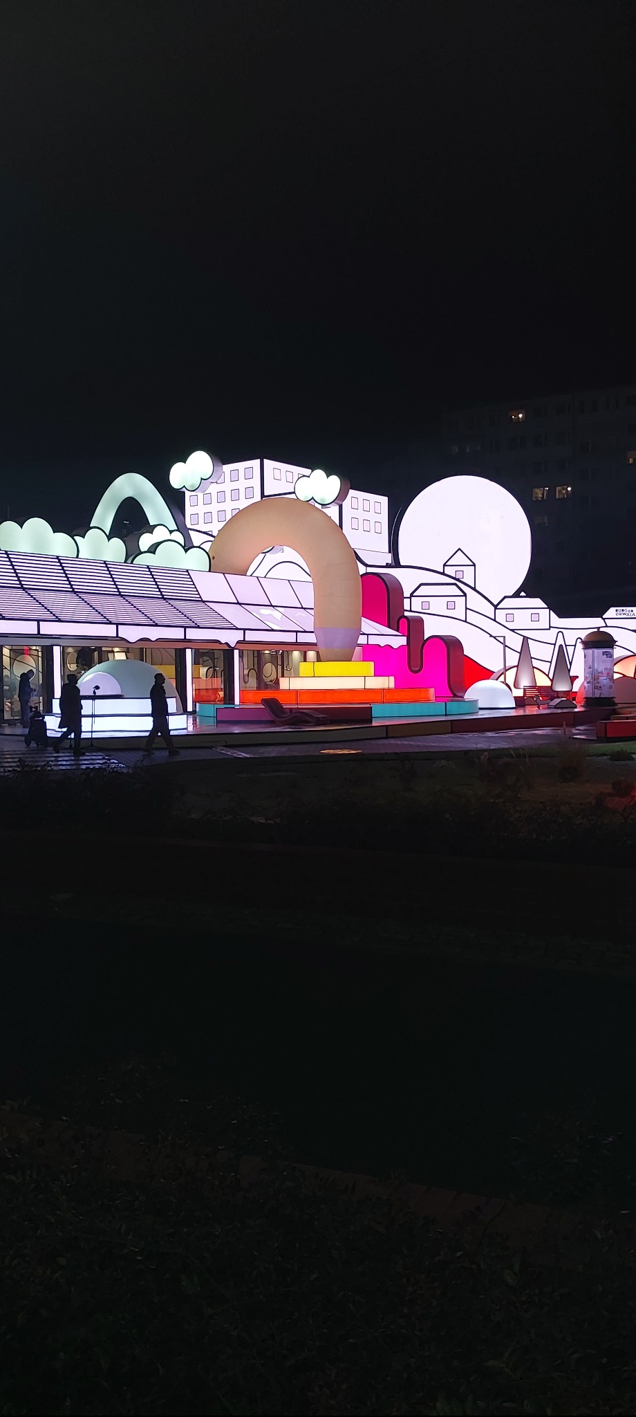 Iluminowana instalacja artystyczna w nocy: kolorowe elementy architektoniczne, sylwetki przechodniów i budynek w tle.