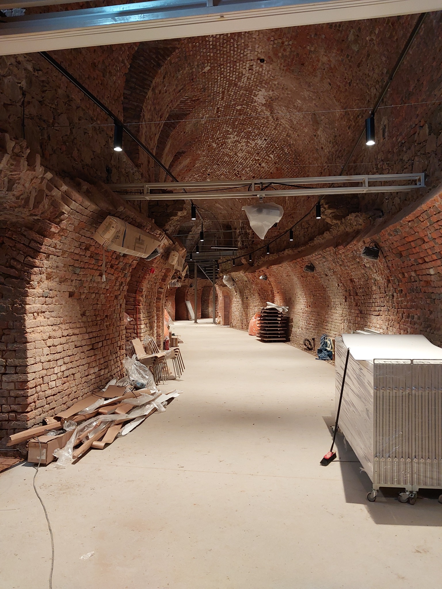 Wnętrze piwnicy z ceglanym sklepieniem łukowym w trakcie prac wykończeniowych, widoczne nowe oświetlenie punktowe na szynoprzewodach, materiały budowlane i przygotowane do montażu składane ścianki...