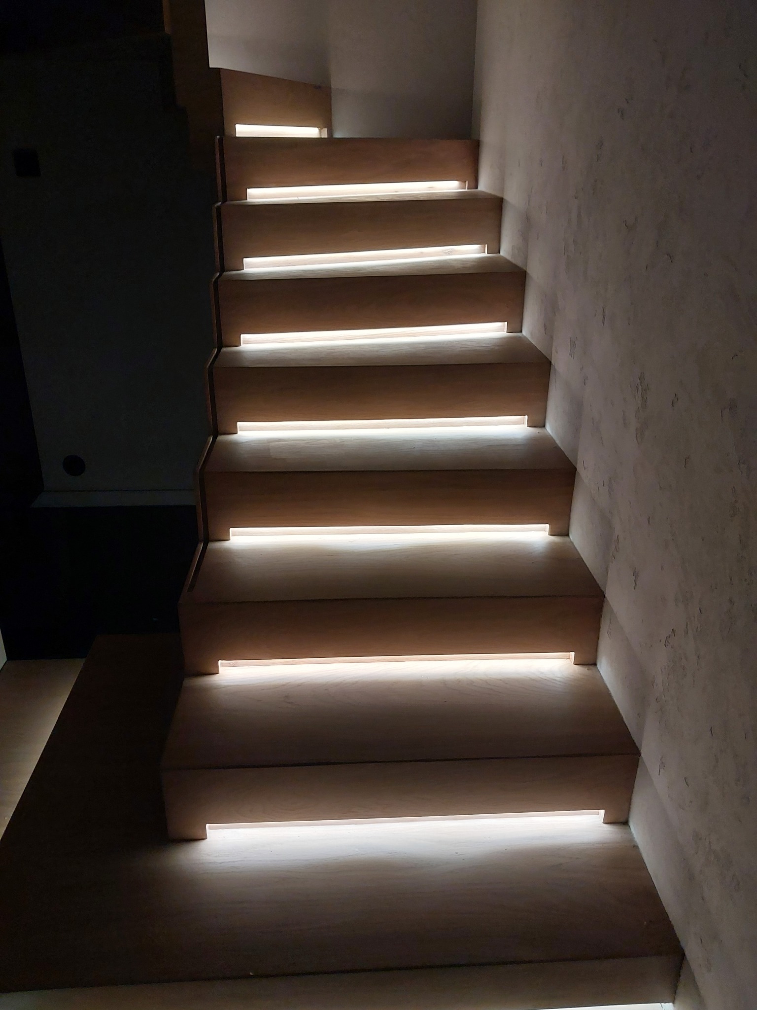 Drewniane schody z podświetleniem LED, widok z dołu, szara ściana z fakturą po prawej stronie, ciemne tło po lewej.