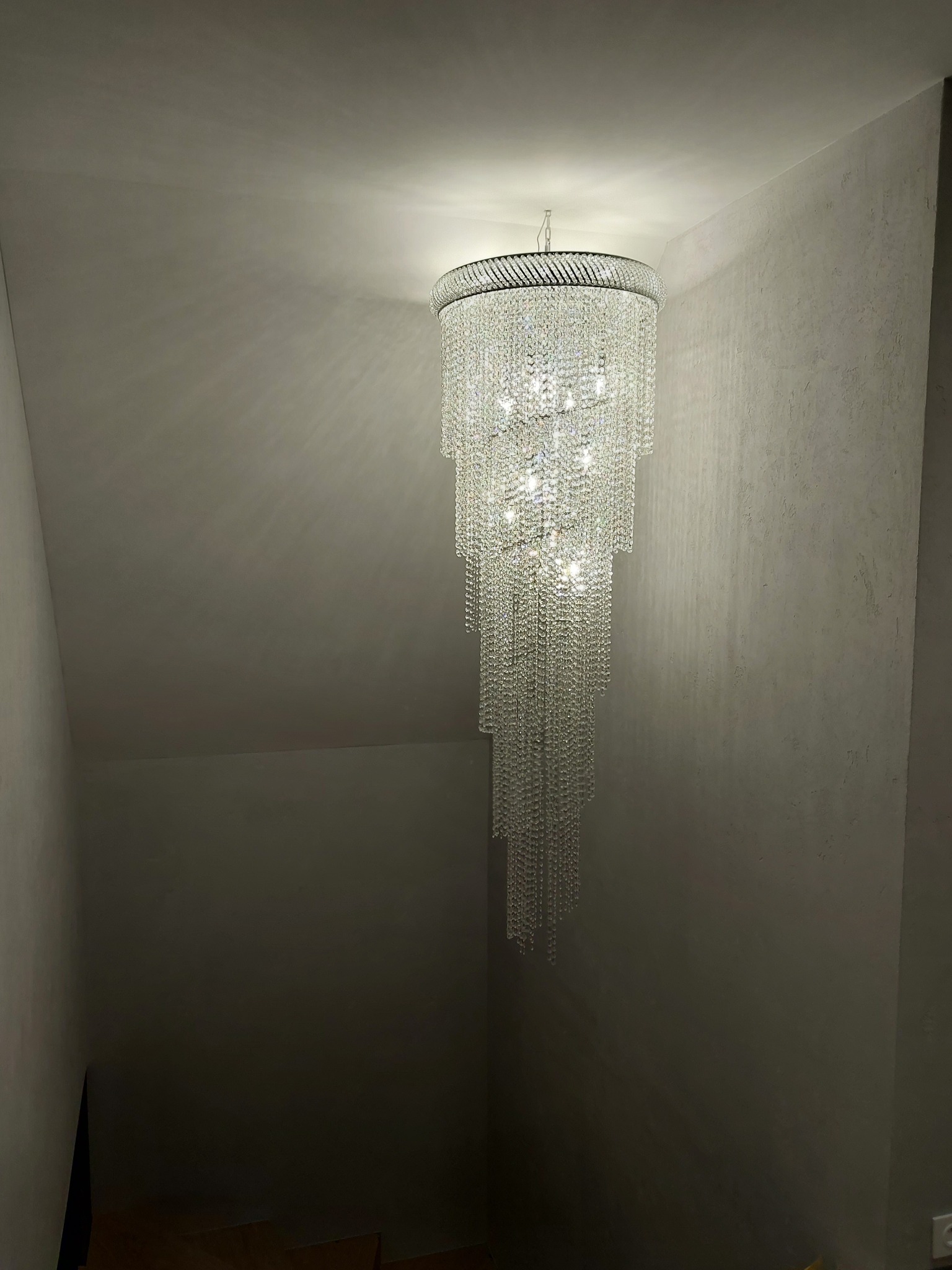 Kryształowy żyrandol kaskadowy z wieloma punktami świetlnymi, zawieszony w klatce schodowej z szarą, teksturowaną ścianą.