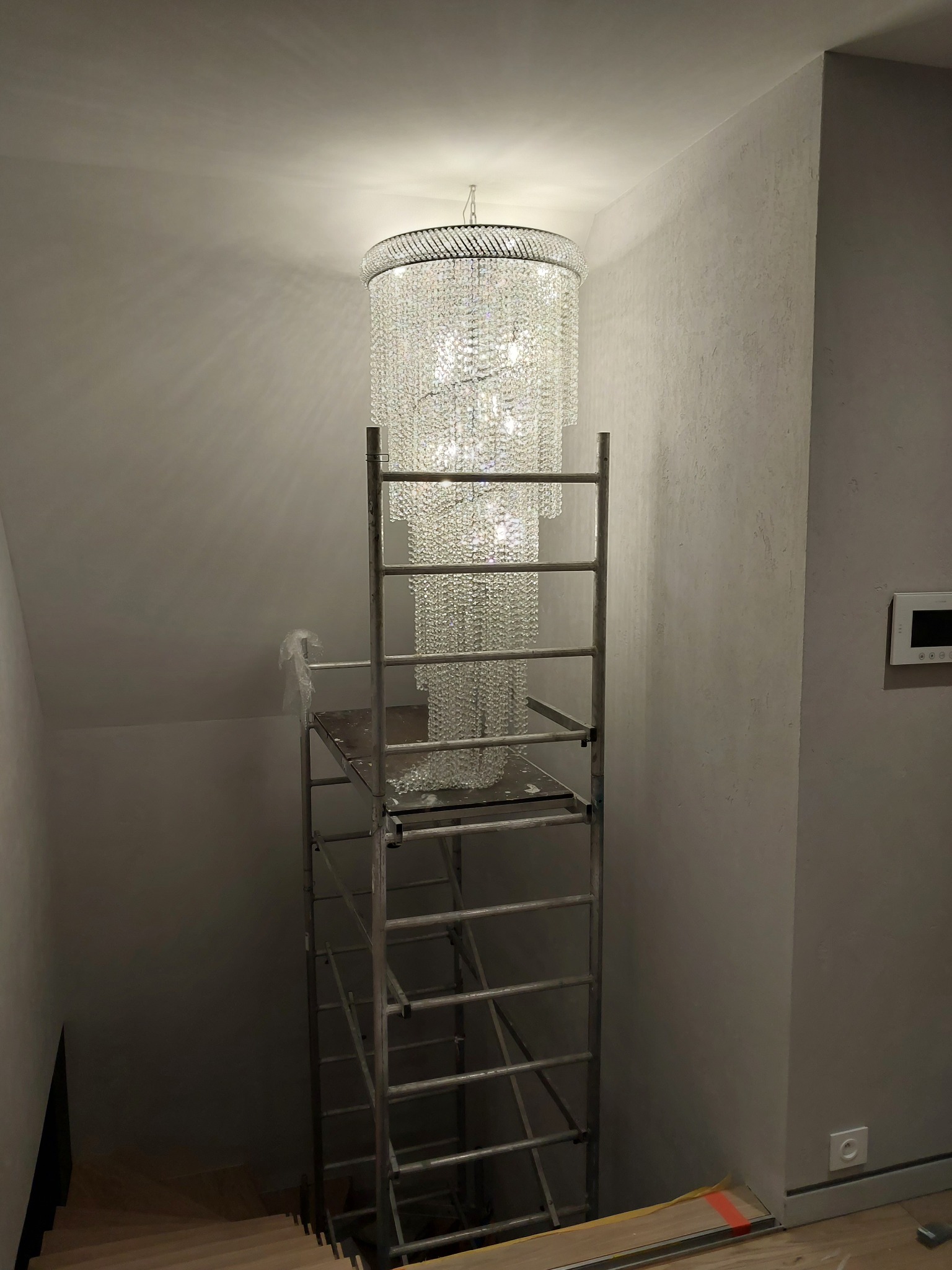 Kryształowy żyrandol zamontowany na klatce schodowej, widoczny ze rusztowania montażowego, ściany w odcieniach szarości z widocznym panelem domofonu.