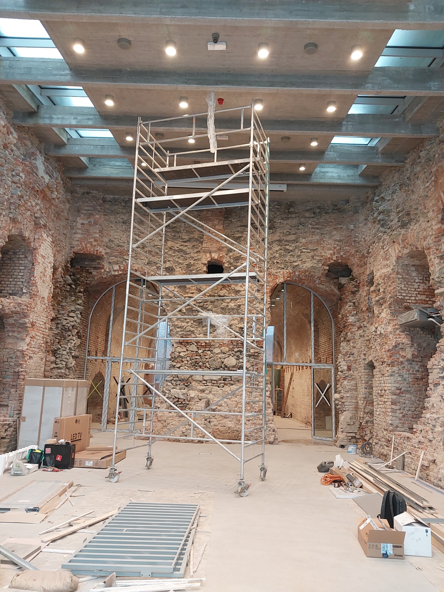 Wnętrze w trakcie remontu z metalowym rusztowaniem na kółkach, widoczne odsłonięte ceglane ściany z łukowymi przejściami, betonowy sufit z oknami dachowymi i zamontowanym oświetleniem punktowym...