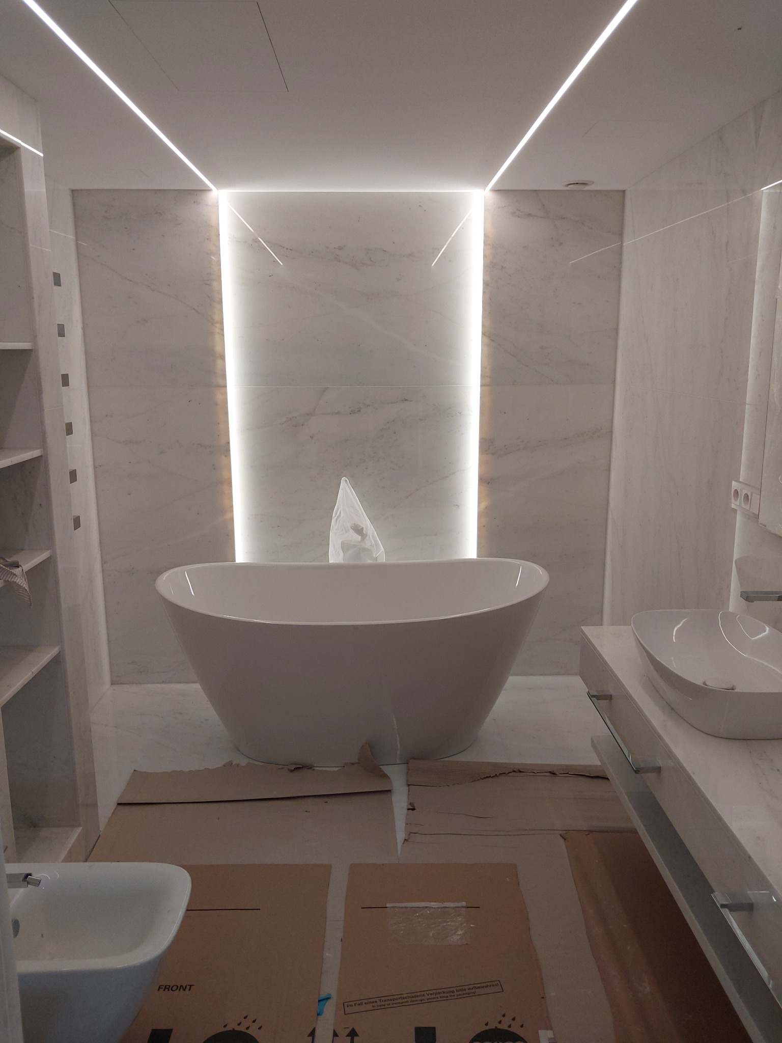 Nowoczesna łazienka w trakcie wykańczania, z wolnostojącą wanną owalną, podświetleniem LED w suficie i na ścianach, zabezpieczona kartonem na podłodze.