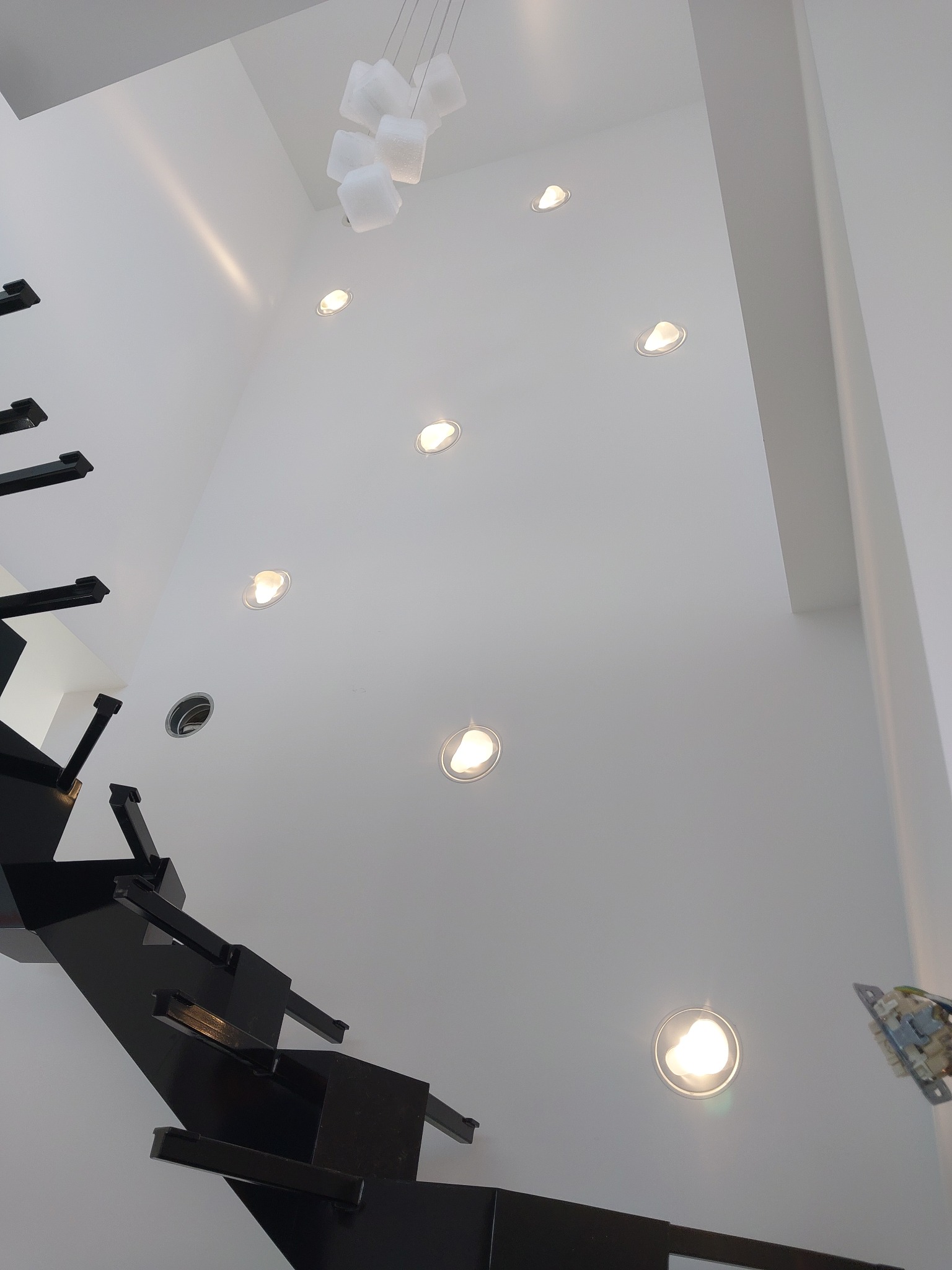 Wysoka ściana z zamontowanymi okrągłymi lampami wpuszczanymi, widok z dołu, obok czarne schody i wisząca lampa z białych kostek.